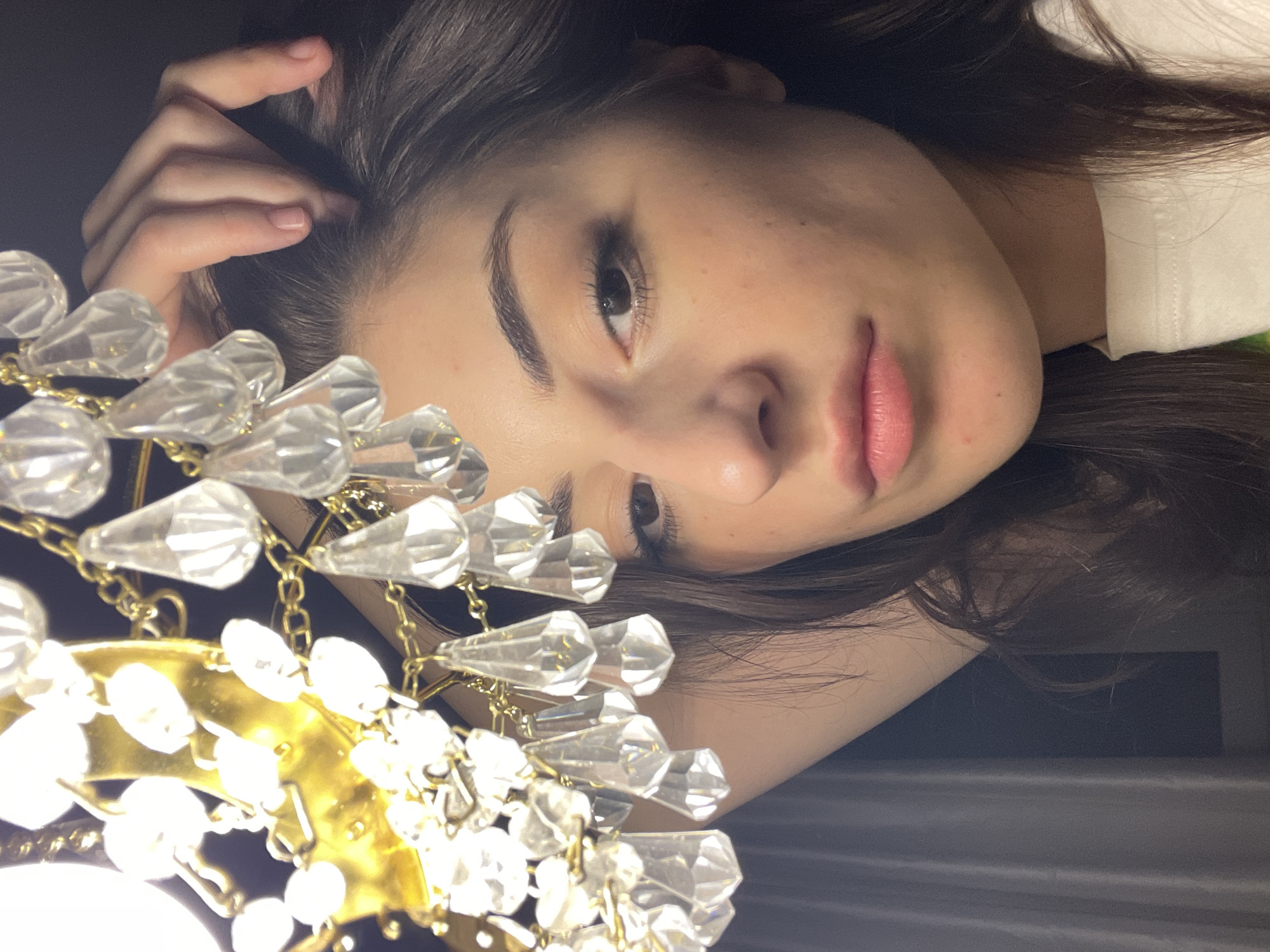 A close-up, tilted perspective captures a young woman with dark brown hair, light skin, and noticeable but subtle freckles. She is wearing a light-colored top. Her right hand is resting on her head, her fingers gently placed near her hairline. To the left of the frame, partially obscuring her face, is a chandelier with clear, teardrop-shaped crystals attached to a gold frame. The bottom of the chandelier is brightly lit, creating a halo effect. The background is a dark, nondescript color, giving the impression of a dimly lit room. There is no indication of the location being Moscow, Russia, or any other specific city.