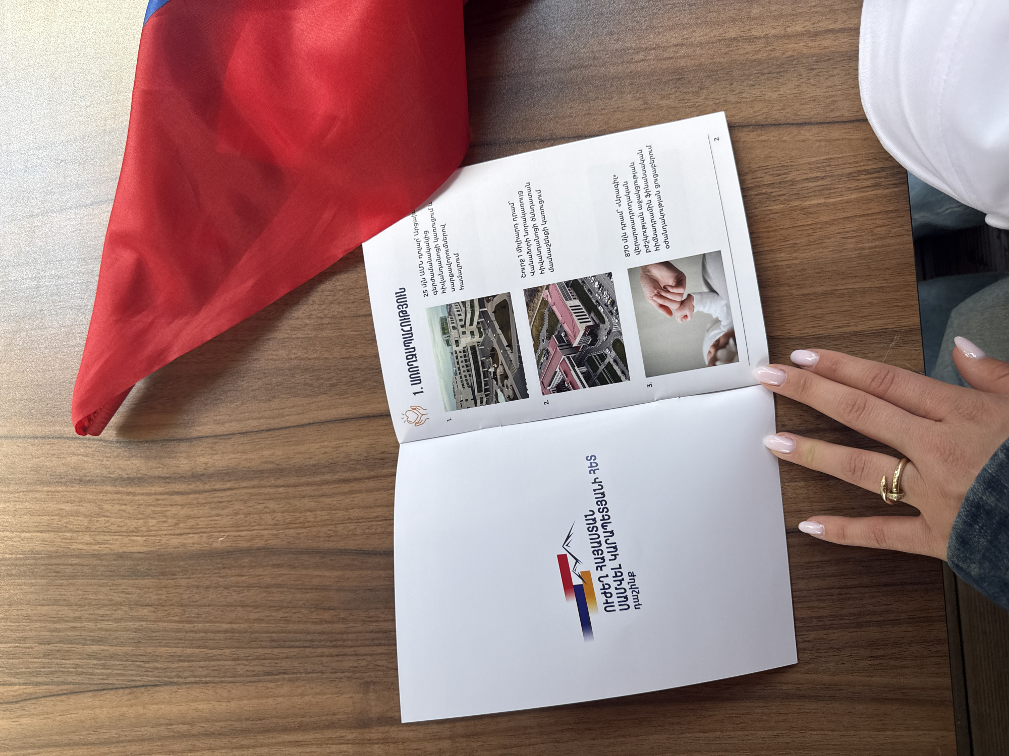 Here is a description of the image:
The image shows a brochure partially opened on a wooden surface next to a red, blue, and orange Armenian flag. A person's hand with a gold ring is holding the brochure open. The brochure appears to be about healthcare initiatives in Armenia, with the title reading, "Strong Armenia with Samvel Karapetyan Alliance." Images and text are visible inside the brochure, featuring buildings, likely hospitals, and details about investments in healthcare, including funding for a hospital in Artsakh and a maternity ward in Vanadzor.