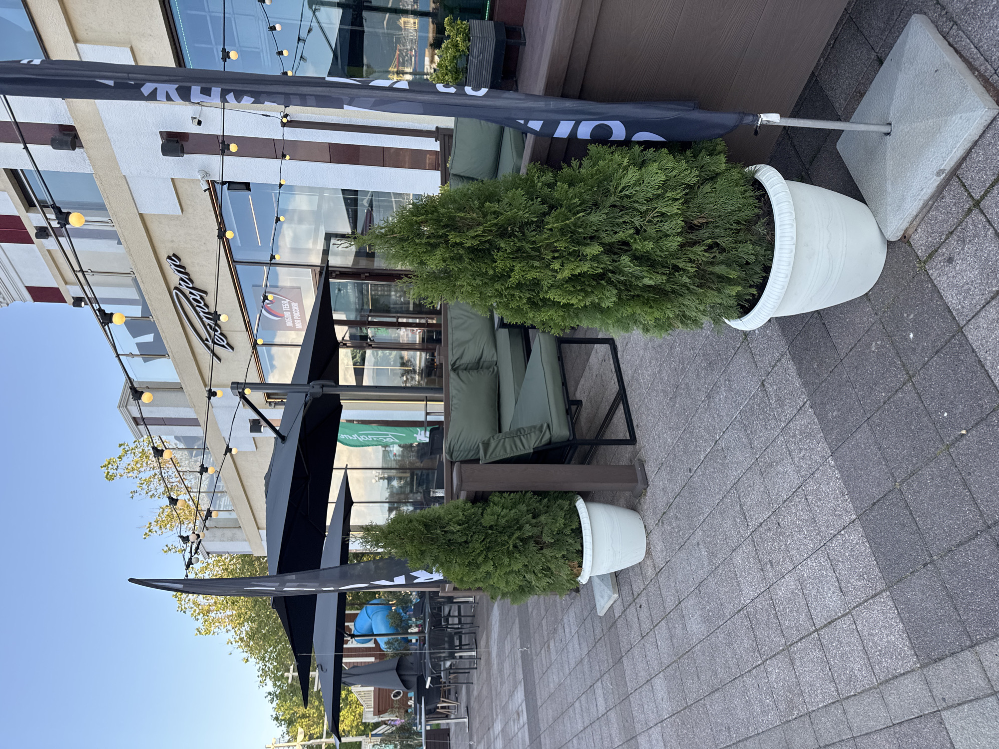 The image depicts an outdoor seating area of a establishment with a modern architectural design, located in Grushovaya Balka, Russia. The foreground features a paved walkway made of gray square tiles, with two large white planters each containing a conical green evergreen tree. To the right of the trees, a dark gray flag stands erect on a metal pole with a concrete base. Behind the planters, a low wooden deck area is visible, furnished with dark green outdoor seating. The establishment itself has a light beige facade with vertical dark red and white stripes, large windows, and an upper-level balcony with a glass railing. A string of yellow globe lights spans across the exterior, casting a warm glow. Trees with yellowing leaves are present in the background, suggesting a temperate season. The overall scene conveys an inviting atmosphere for outdoor dining or relaxation.