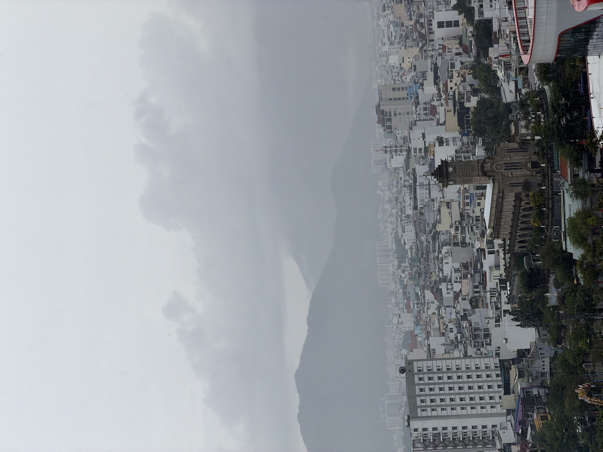 An aerial view of Nha Trang Ward, Vietnam, under a cloudy sky, reveals a densely packed urban landscape abutting a prominent mountain. Buildings in varying shades of white and gray intermingle with patches of green trees. A tall stone church with a clock tower stands out among the buildings. In the top right corner, a red and white structure is partially visible. The mountain appears hazy due to the weather conditions.