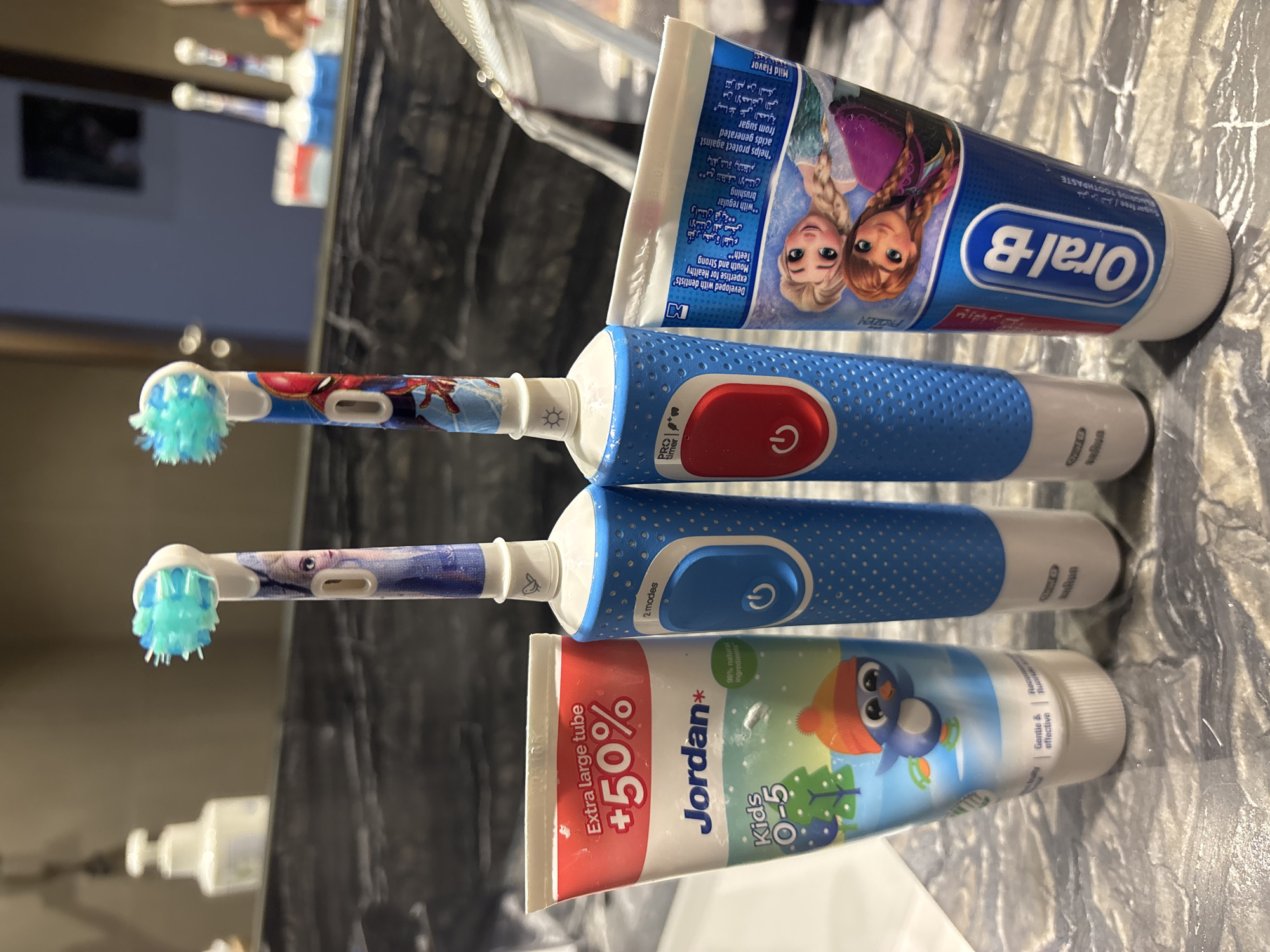 The image displays a close-up, eye-level shot of two children's electric toothbrushes and two tubes of toothpaste arranged on a marbled surface, likely a bathroom countertop. The toothbrushes are blue and feature characters from Disney's Frozen on one and Marvel's Spider-Man on the other. Both toothbrushes have white brush heads with blue bristles and a single power button. To the left, a tube of "Jordan" brand toothpaste for kids aged 0-5 is visible, decorated with a cartoon penguin. To the right, an "Oral-B" toothpaste tube featuring characters from Frozen is partially obscured by the toothbrush. The background is blurred, showing a reflection of a bathroom sink and a dispenser. The scene suggests a domestic bathroom setting. The image does not contain any information about the city of Dubai, United Arab Emirates.
