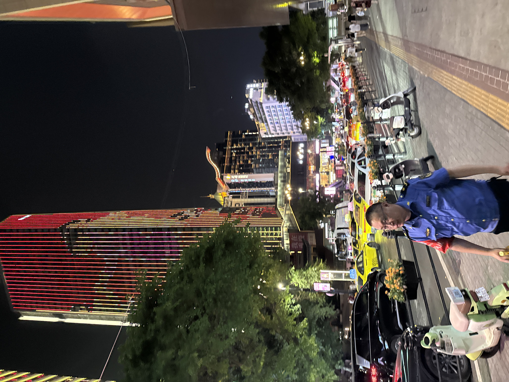 A man in a blue uniform with red and blue sleeves walks on a sidewalk at night in Yuzhong District, China. He is wearing glasses and holding a yellow object in his right hand. To his left, a light green electric scooter is parked. In the background, a street is lined with cars, including several yellow taxis and a black SUV. Tall buildings, illuminated with red and yellow lights, dominate the skyline. Trees with green foliage are interspersed between the buildings and the street. The sidewalk has a textured yellow and brown tactile paving. Several other people are visible walking in the distance.