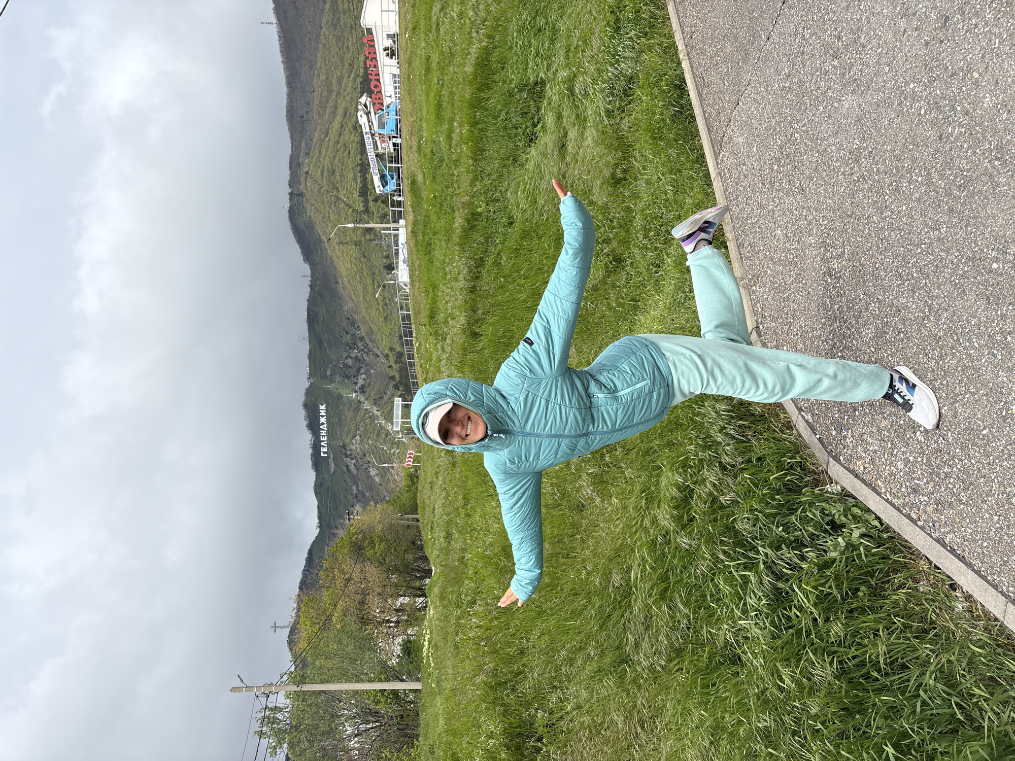 A woman in a light blue tracksuit and white cap stands with arms outstretched in a field of tall grass next to a gray sidewalk in Gelendzhik, Russia. Behind her, a hillside is visible with the name of the city, "ГЕЛЕНДЖИК" (Gelendzhik), written in large white letters. The sky is overcast. The hillside is green with trees and what appears to be a cross on the hilltop. In the distance, there is a large building or structure with red and blue accents.