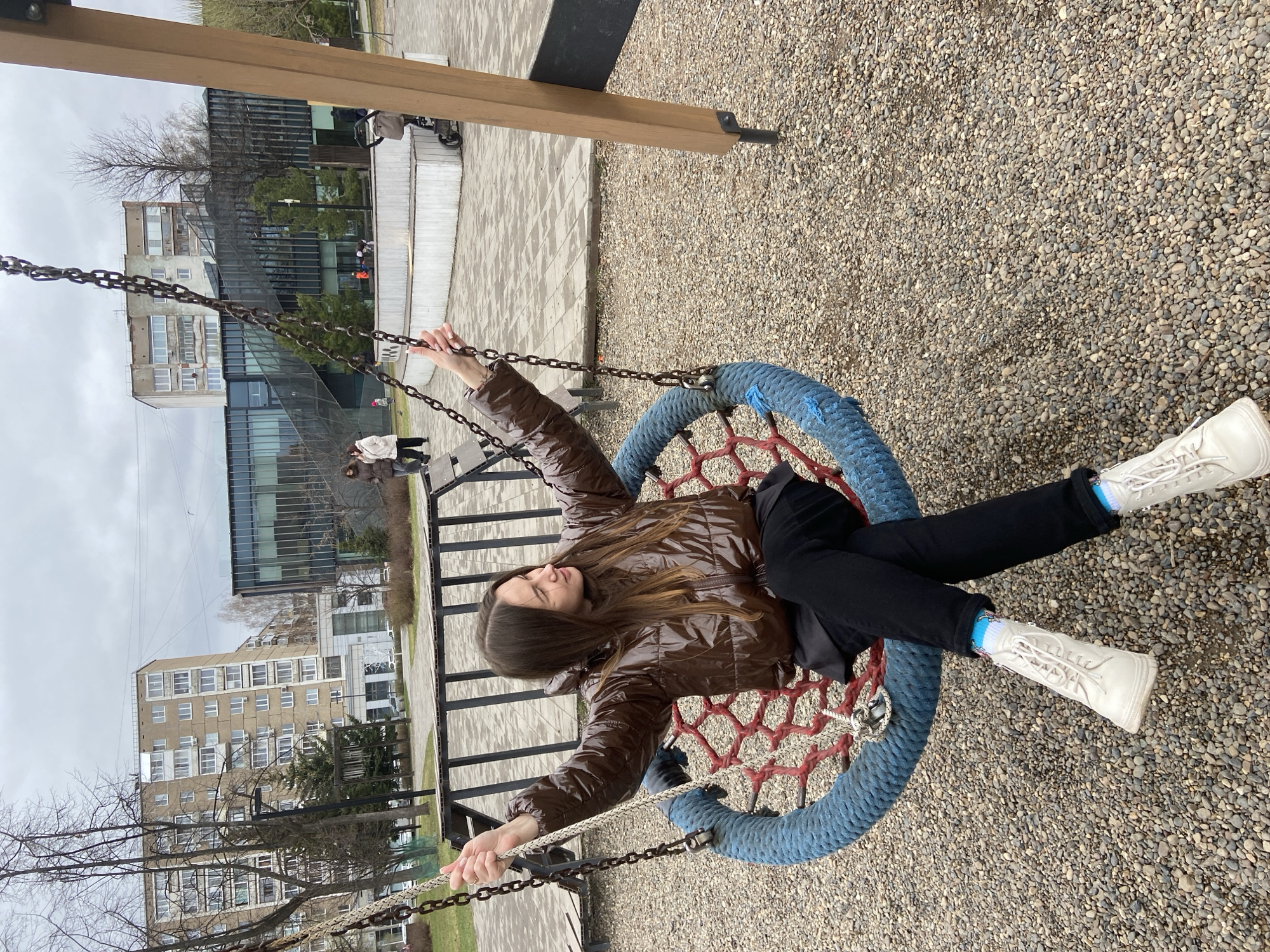 A young woman is shown relaxing on a large round "nest" swing in an urban playground or park, set in Izhevsk, Russia. She has long dark brown hair and is wearing a shiny brown puffer jacket, black pants, and white high-top sneakers with light blue and pink patterned socks. She lies on her back in the swing, holding onto the metal chains, with a calm or thoughtful expression as she looks upwards.
The swing features a blue padded rim and a red net base. The ground beneath the swing and to the right is covered in light-colored gravel, while paved areas are visible further back. The background features a mix of architecture: a multi-story residential building with numerous windows and balconies, and a more modern building with a dark glass facade. Several bare trees indicate the time of year is likely late winter or early spring. The sky is overcast and grey, suggesting a cloudy daytime. A few other people are faintly visible in the background, including one person seated on a bench and another with a stroller. No discernible text is visible.