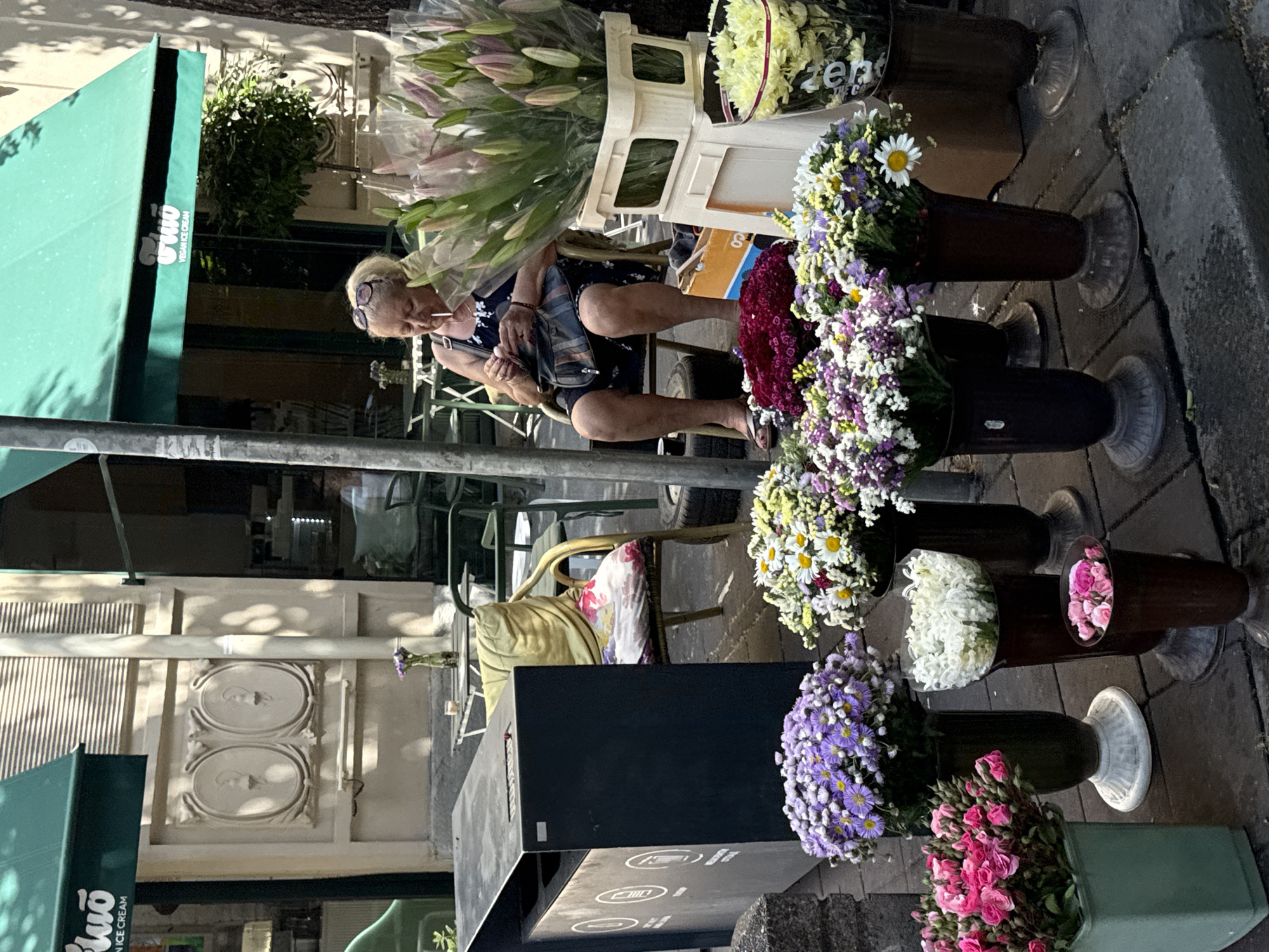 The image shows a woman smoking a cigarette while sitting on a cart filled with flowers, next to a flower stand. The flower stand displays various colorful flower arrangements in vases, including pink roses, purple asters, white daisies, and a mix of white and yellow flowers. There are also bundles of lilies in plastic wrap behind the woman. The scene appears to be on a sidewalk in Belgrade, Serbia, indicated by the "Bio Vegan Ice Cream" awning in the background. Behind the stand are tables and chairs, suggesting an outdoor cafe. The building behind the cafe has ornate architectural details.