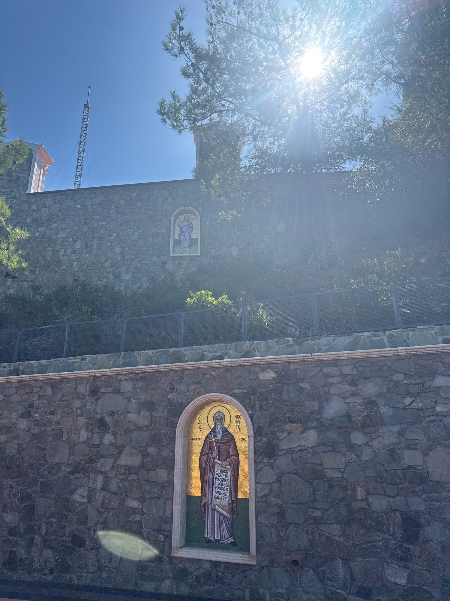 The image shows a stone wall with two mosaics of religious figures. The larger mosaic, located in the foreground, depicts a bearded man with a halo, wearing a purple robe and holding a scroll. The mosaic is framed by a decorative border. The wall itself is constructed from irregular, dark-colored stones. Above this wall, a second, smaller mosaic of a robed figure is visible on another stone wall. In the background, a tall metal structure, possibly a tower or antenna, is visible against a clear blue sky. Sunlight streams through tree branches, creating a lens flare effect. The scene appears to be outdoors, with vegetation visible behind the walls. The location is identified as Mylikouri, Cyprus.