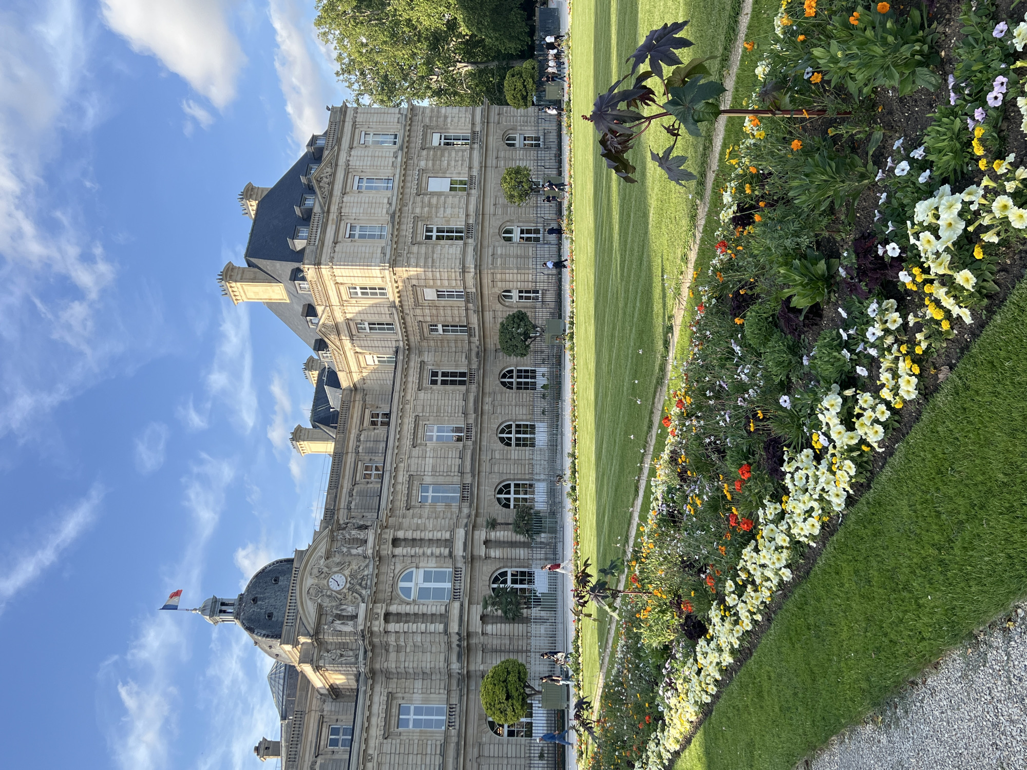 The image depicts a scene in the Luxembourg Garden in Paris, France. In the background, a grand building with classical architecture, featuring a dome topped with the French flag and a clock, stands prominently. The building is constructed of light-colored stone and has numerous windows. In front of the building, a meticulously maintained green lawn is visible, bordered by a vibrant flowerbed filled with a variety of colorful blossoms, including white, yellow, orange, and red flowers. Several trees are strategically placed around the lawn. In the middle ground, a few individuals are walking on a path behind a metal fence, and a small dog is also visible on the lawn. The sky above is blue with scattered white clouds, suggesting a pleasant day. The foreground is dominated by the flowerbed and a gravel path.