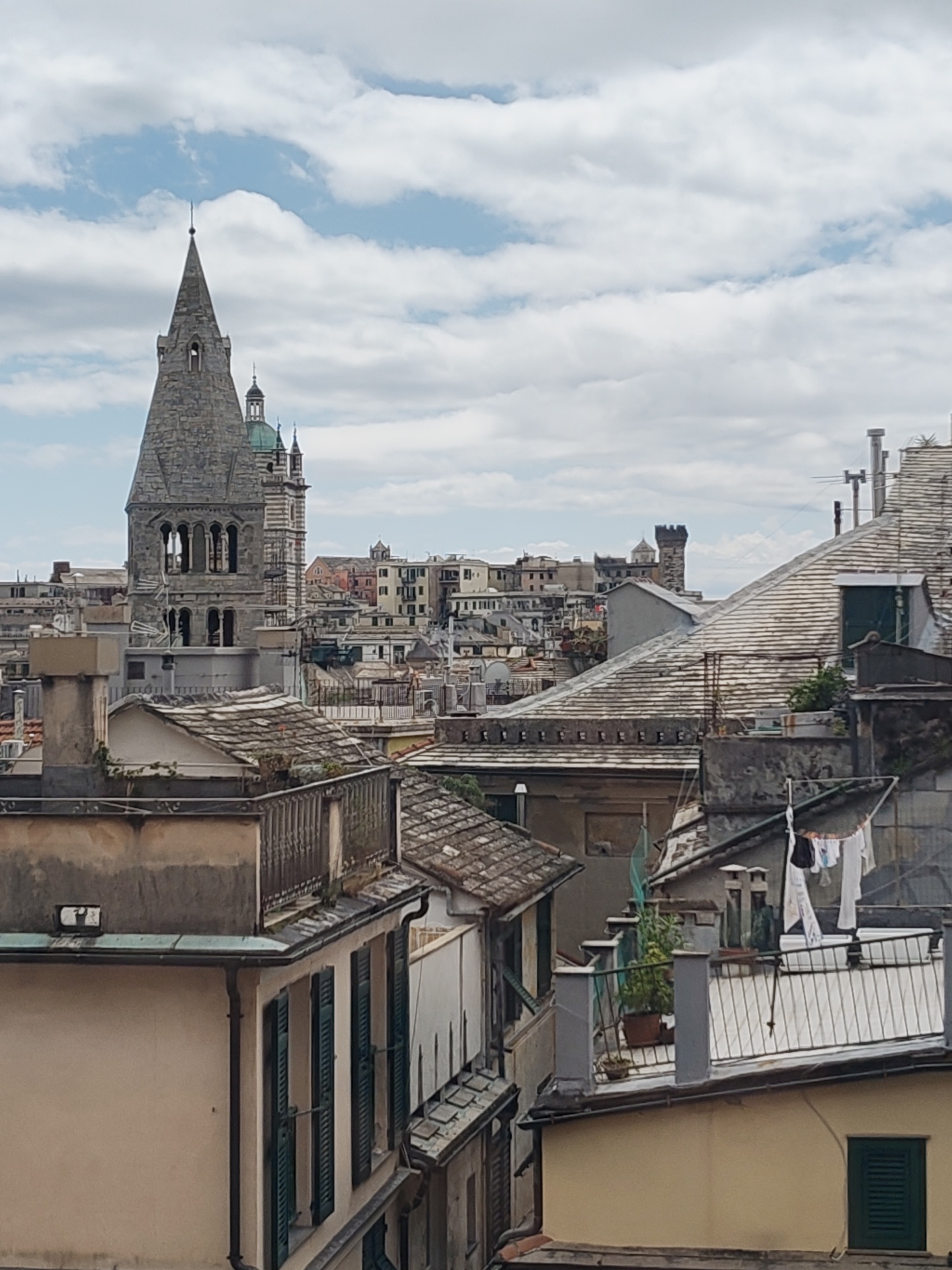 The image depicts an urban landscape in Genoa, Italy, characterized by a dense arrangement of buildings with varying heights and architectural styles. In the background, a prominent stone tower with a pointed spire rises above the surrounding structures. The foreground features rooftops of adjacent buildings with tile patterns, chimneys, and vegetation. Laundry hangs on a clothesline on one of the rooftops. The sky is overcast with clouds.