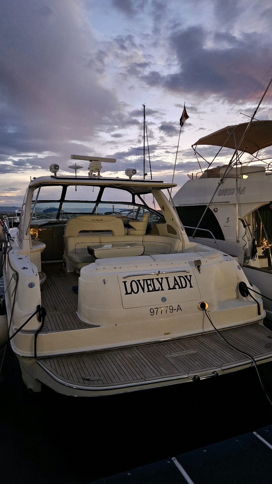 A rear view of the motor yacht "Lovely Lady" is shown docked in the marina of Općina Tučepi, Croatia, during twilight. The yacht is white with a wooden deck and a tan canopy. The name "Lovely Lady" is written in black lettering on the stern, above the registration number "97779-A". Another white motor yacht with the registration number "91529 VA" is docked to the right. The sky is cloudy with shades of purple and orange, indicating sunset or sunrise. The water is dark, reflecting the sky.