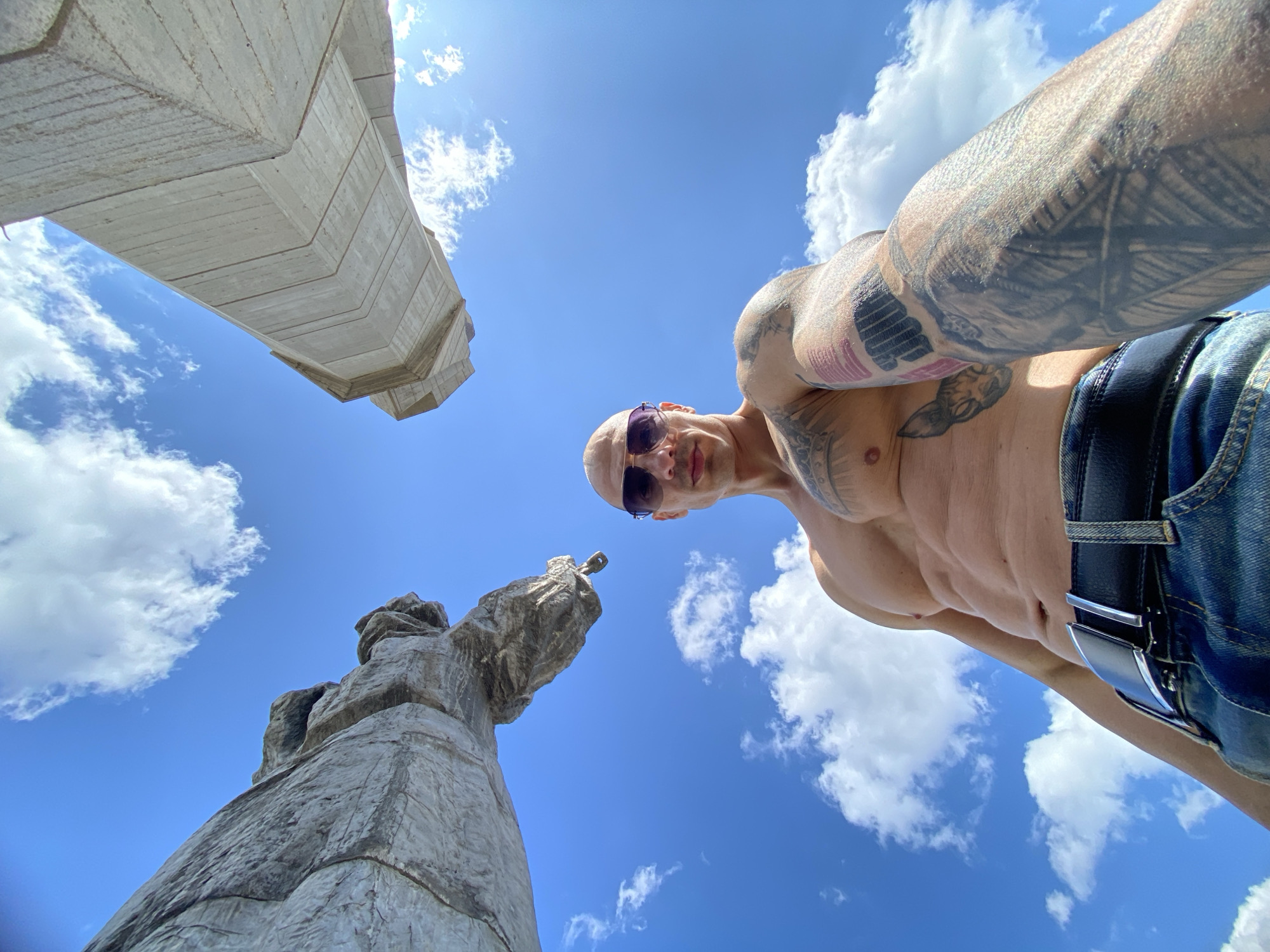 A low-angle shot reveals a man wearing sunglasses, shirtless, and denim jeans with a black belt, positioned against a backdrop of blue sky with scattered clouds. His arms are heavily tattooed. In the foreground, an abstract concrete structure is visible on the left. Further back on the left is the towering monument “Aleksandrovski Trakiyski Kurgan” located in Pleven, Bulgaria. The perspective is from below, looking up at the man and the monument.