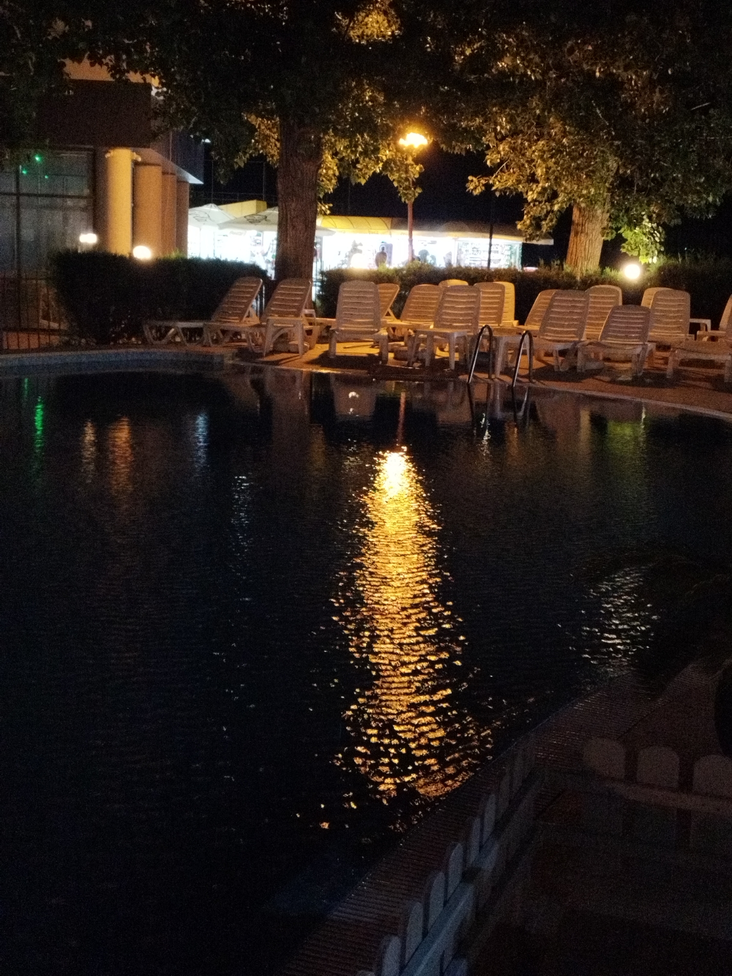 This is a low-light, outdoor photograph of a swimming pool at night, likely at a hotel or resort. In the background, there is a building with illuminated columns and windows, as well as several white lounge chairs arranged around the pool. Trees with lush foliage are visible, casting shadows and reflecting light from a streetlamp onto the water. The surface of the pool is dark, with shimmering reflections of the lights from the surrounding area, creating an atmospheric effect. The image appears to have been taken in Constanța, Romania, given the reference.