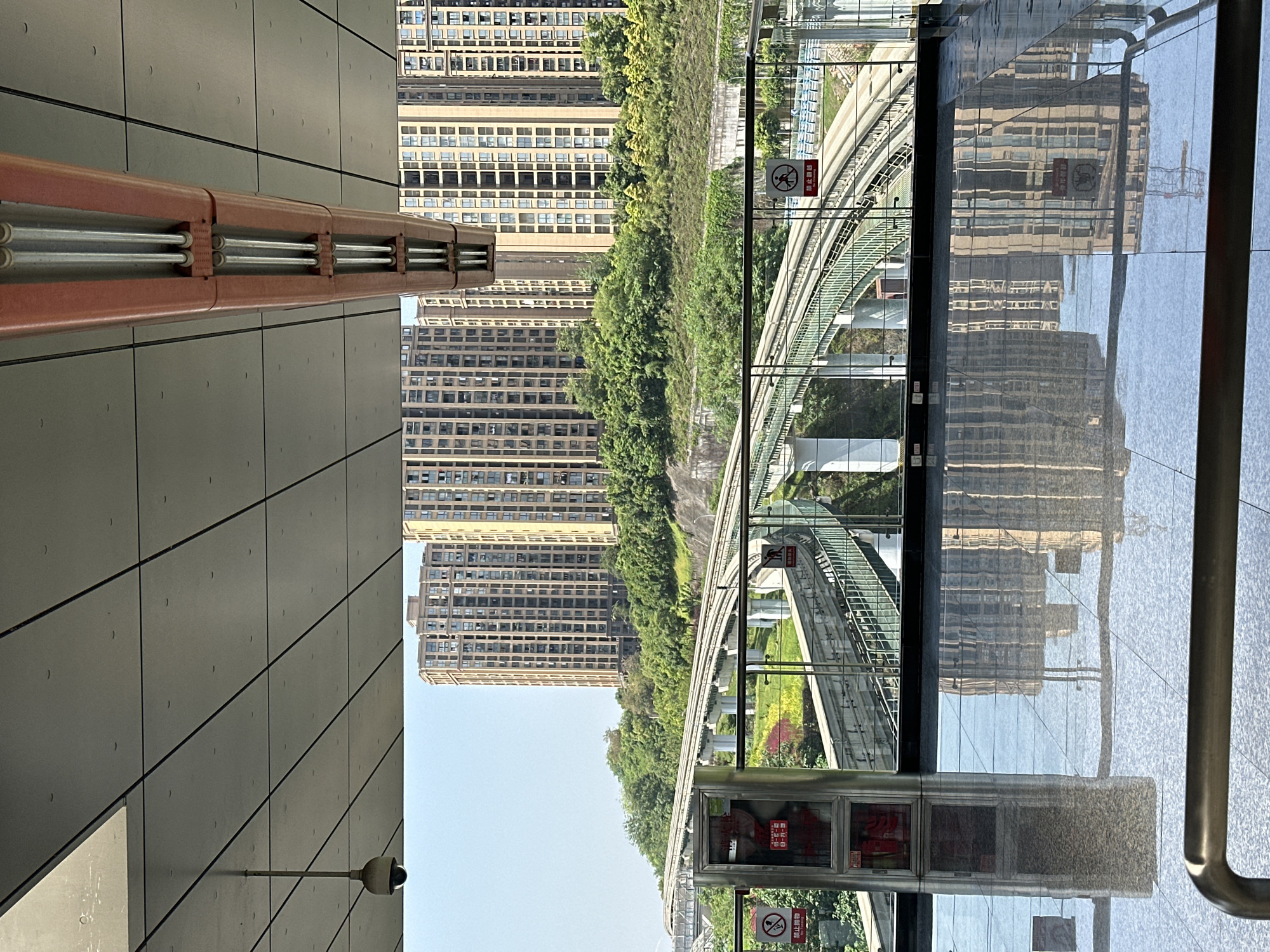 The image shows a scene in Jiansheng, China, featuring a building complex adjacent to an elevated monorail track. The building, consisting of multiple towers, is viewed through a glass partition reflecting the cityscape. The monorail track is supported by white pillars, weaving through greenery, suggesting an urban integration with nature. A gray tiled wall is seen with a CCTV camera. A prohibition sign is visible on the glass partition. A fire safety box with a red warning label is present.