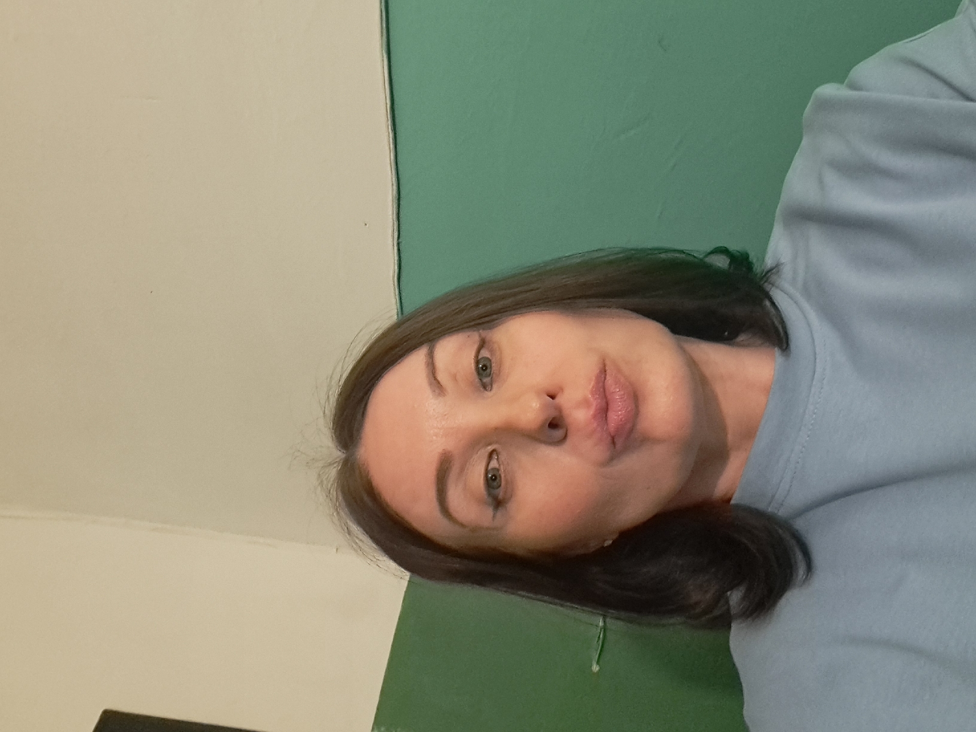 A medium shot shows a woman with dark hair and fair skin posing for a selfie against a teal and off-white wall. The woman is looking towards the camera with pursed lips and her eyes are open and looking forward. She is wearing a light blue sweatshirt. The background consists of a teal lower half and an off-white upper half, divided by a thin horizontal line. The lighting is dim and artificial, casting a slight shadow on the woman's face. There are no visible landmarks or identifying features that would indicate a specific location in Irkutsk, Russia.