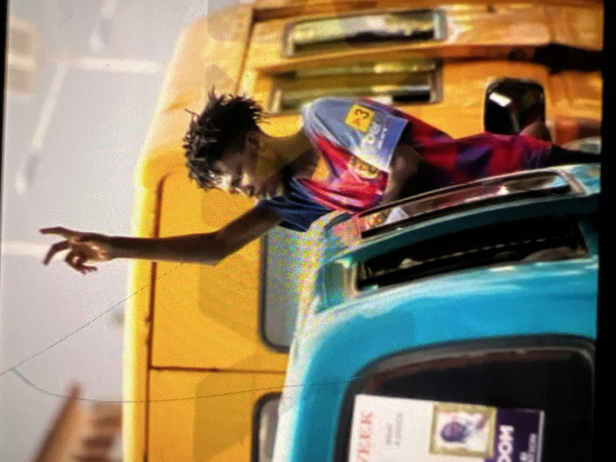 The image shows a young man with dreadlocks, wearing a red, blue, and purple striped shirt, standing with his right arm raised, hand open. He appears to be in an urban environment, possibly a street in Lagos, Nigeria, given the presence of a yellow commercial bus, commonly known as a danfo, which is a prevalent mode of transport in the city. A teal-colored vehicle is positioned in the foreground, partially obscuring the view of the danfo. A sign is visible on the teal vehicle with the word "WEEK" and a framed picture of a person. The man's expression is neutral, and his posture suggests he might be hailing a vehicle or signaling to someone. The lighting indicates it is daytime.
