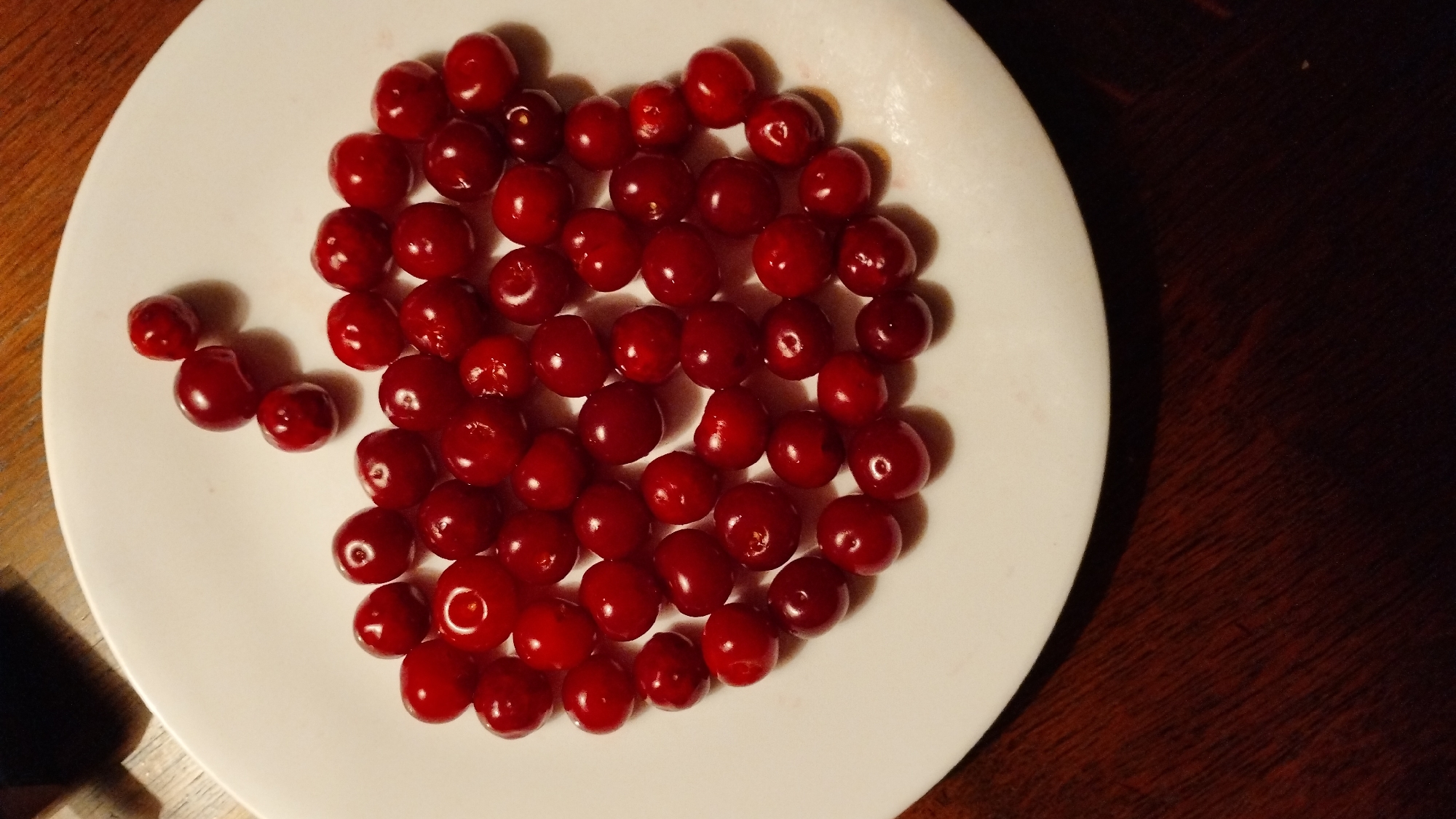 A white plate filled with dark red cherries sits on a dark wood table. The cherries are scattered across the plate in a loose pile, with some overlapping. The lighting is dim, casting shadows on the table and highlighting the glossy surface of the cherries. The plate is positioned in the center of the frame, with the table surface visible around it.