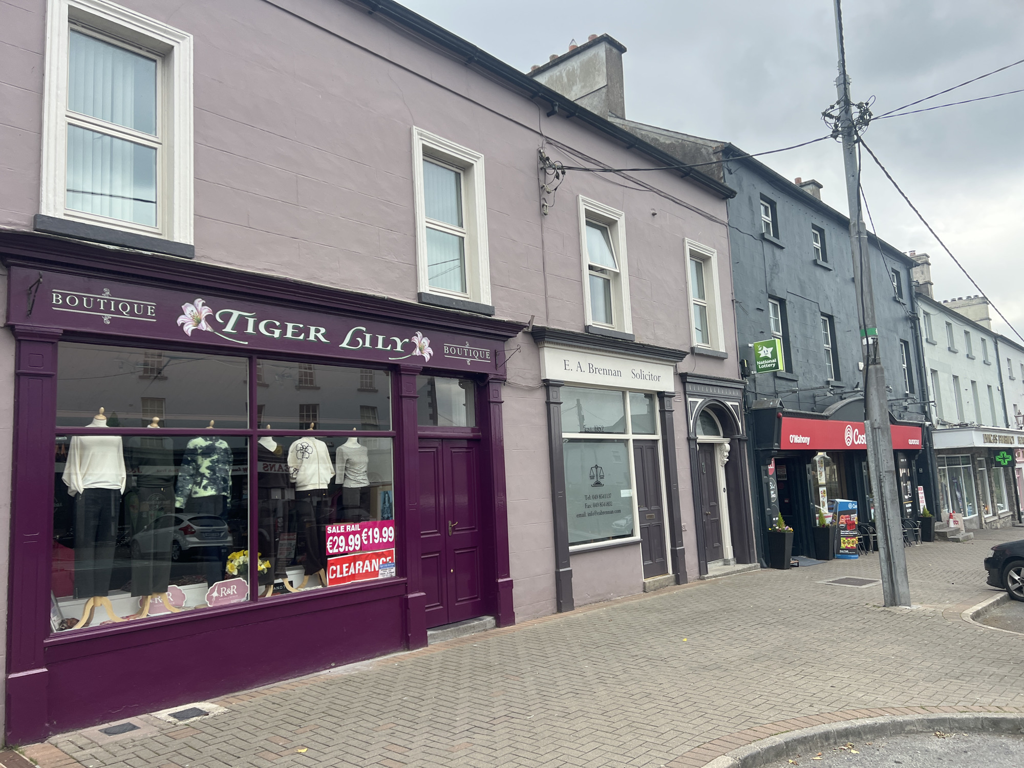 The image shows a street scene in an unknown city. On the left, there is a purple storefront with a sign that reads "BOUTIQUE TIGER LILY". The shop window displays mannequins dressed in clothing. To the right of the boutique, there is a light purple building with a sign for "E.A. Brennan Solicitor". Further down the street, there are other businesses with various signs, including "O'Mahony" and "Costcutters". The street is paved with cobblestones, and there is a lamppost with many wires on the right side of the image. The sky is overcast.