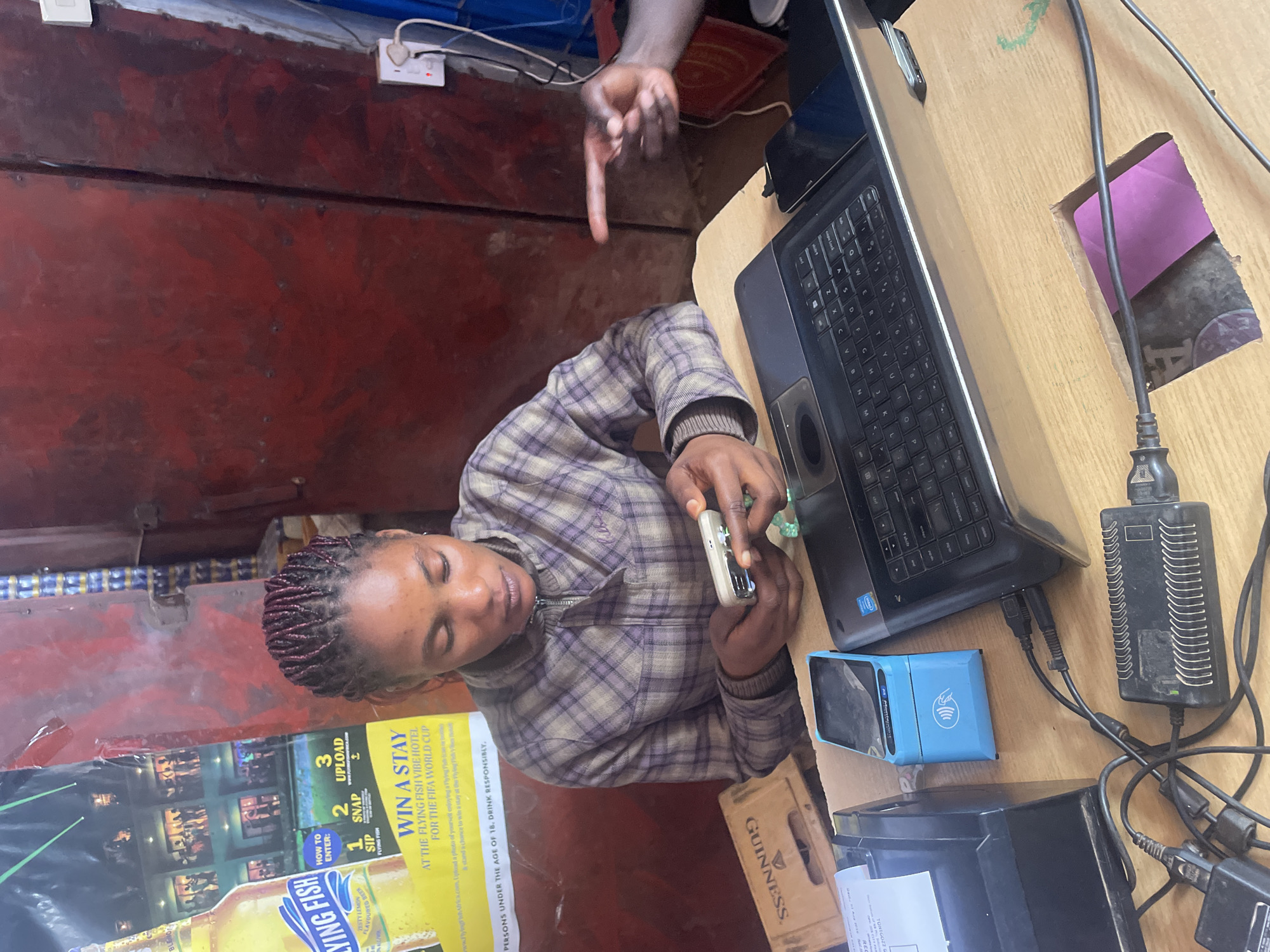 In Gura Top, Nigeria, a woman with braided hair sits at a wooden counter, engrossed in her mobile phone. She wears a purple and grey plaid jacket. In front of her are a laptop, a receipt printer, a blue card reader, and a Guinness crate. A poster advertising Flying Fish beer and a competition is visible to her left. Another person's hand points towards the woman from the right side of the frame. The background features a distressed red metal door and electrical outlets with cords.