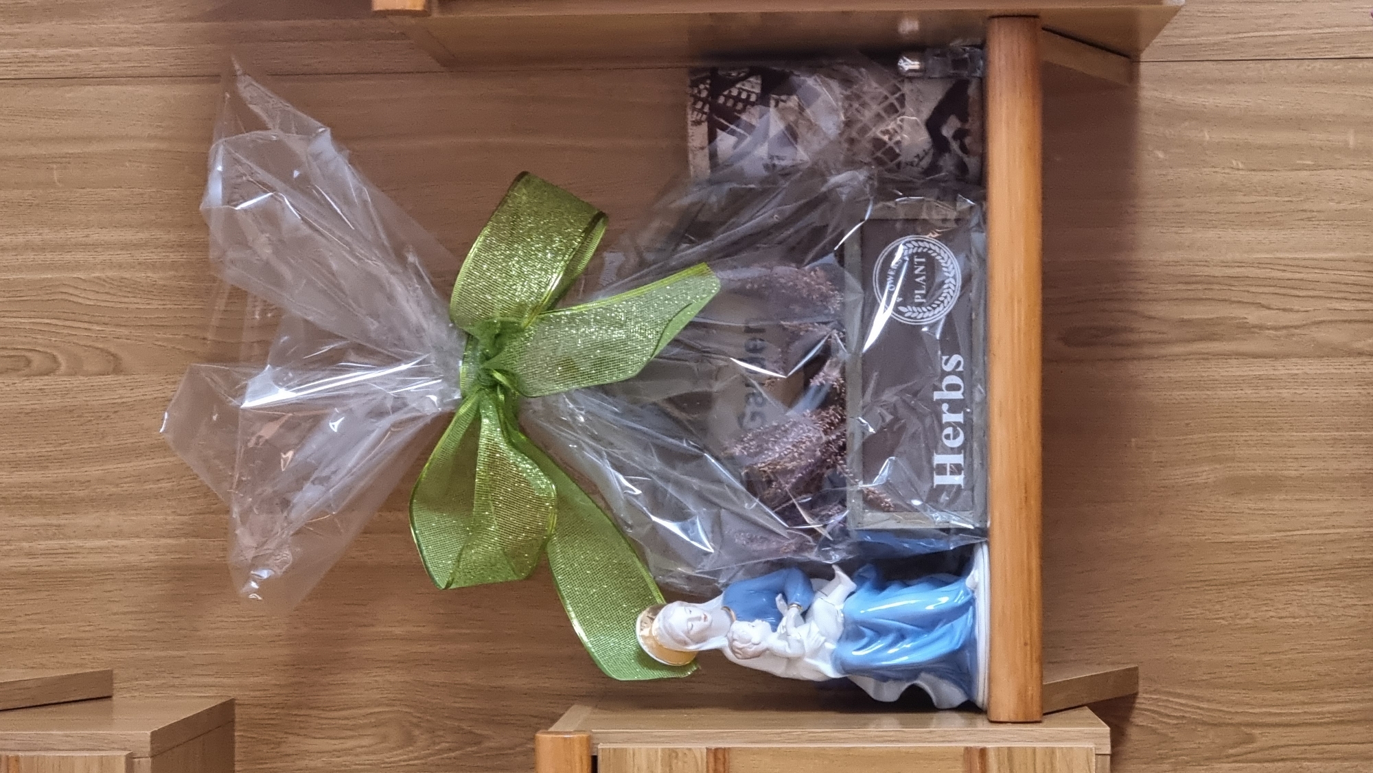 A gift basket is displayed on a wooden shelf against a wood-paneled wall. The basket is wrapped in clear cellophane and tied with a sparkly green ribbon. Inside the basket, a figurine of Mary holding the infant Jesus is visible on the left. To the right of the figurine, there are various items within the cellophane, including what appears to be a small wooden box labeled "Herbs," and possibly some dried herbs or potpourri. Further to the right, there are cylindrical objects, one of which has a patterned surface. The overall presentation suggests a thematic gift, possibly related to gardening or herbs, with a religious element from the figurine. The image does not provide any specific information about its location in Sant Joan Les Fonts, Spain.
