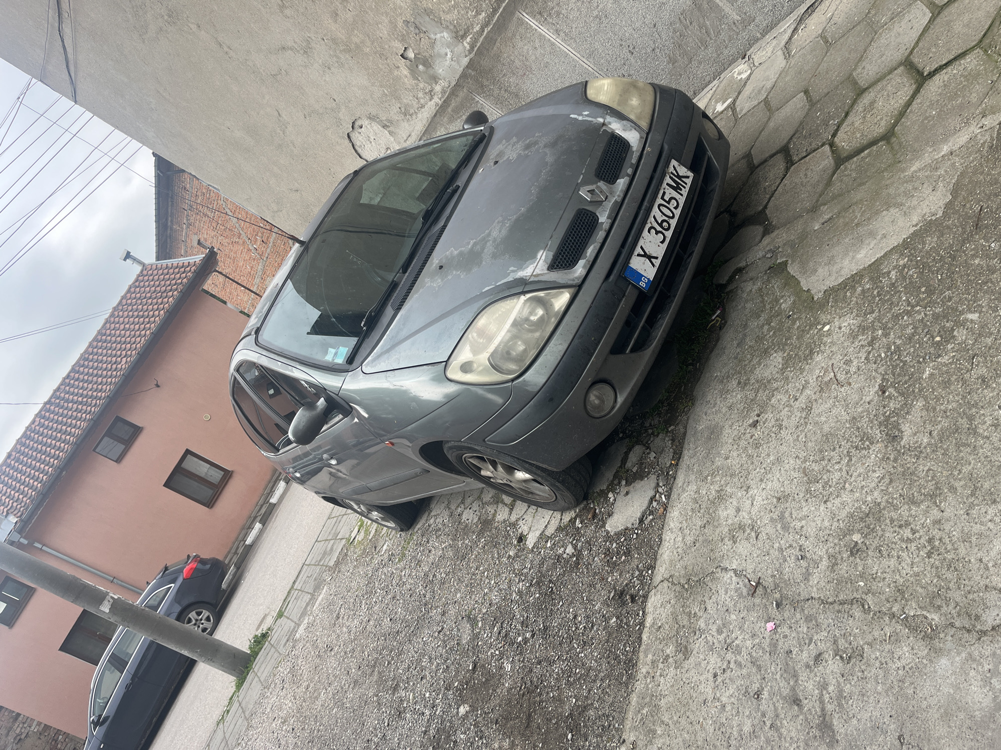 A gray Renault Scenic, license plate X 3605 MK, is parked on a gravel and concrete surface next to a sidewalk made of interlocking concrete blocks. The car's paint is faded. To the left, a pink building with a tiled roof and several windows is visible, as is a dark-colored car parked nearby. A utility pole is positioned in front of the building. The location appears to be an alleyway or narrow street in a residential area. Based on the license plate, the location is likely Bulgaria. No people are visible.