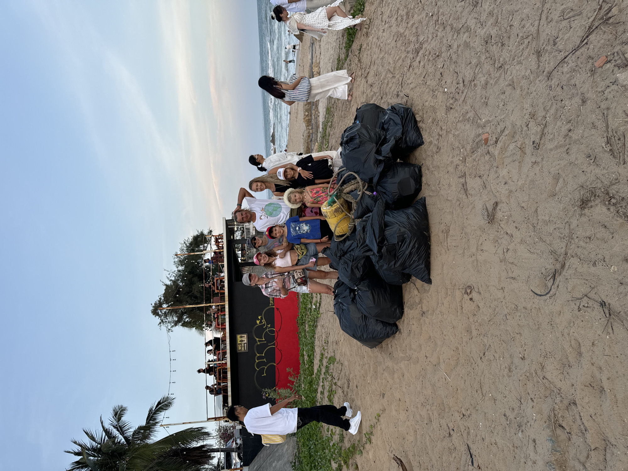 On a sandy beach in Mũi Né, Vietnam, a group of approximately 15 people of varying ages, including adults and children, pose for a photo next to a pile of six black trash bags filled with refuse collected from a beach cleanup. The group includes individuals with a mix of fair and darker skin tones, and attire ranges from casual t-shirts and shorts to dresses and hats.
The trash bags are centrally located in the scene, serving as a visual testament to the activity. Behind the group is a dark-colored building with an open-air seating area filled with people; the building features graffiti on a red painted section.
In the foreground, a young man with dark hair wears a white t-shirt, black pants, and white sneakers; he looks back at the main group while holding his hands out to each side. A palm tree is visible in the lower left corner.
In the background, the ocean meets the sky along the horizon. Closer to the water, two women in dresses stand at the water's edge. The sky is a pale blue, indicative of early morning or late afternoon light. The sand of the beach is light brown, showing the texture of the grains.