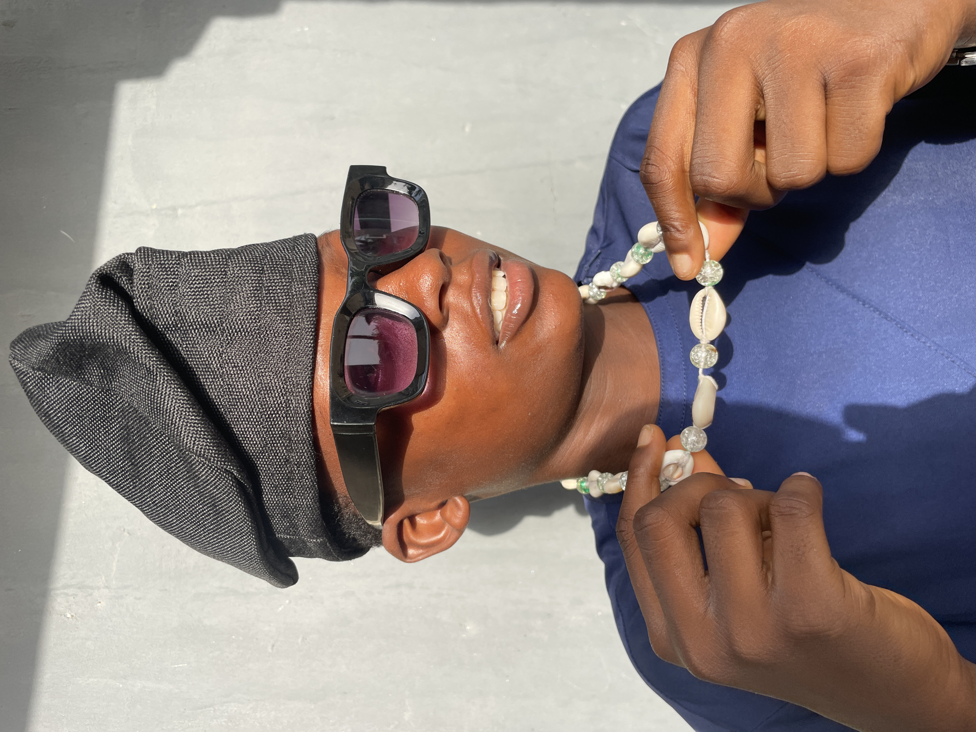 An overhead shot captures a person with dark skin lying down or leaning back, looking upwards, in an outdoor setting in Badagry, Nigeria. The image is rotated 90 degrees, presenting the subject's head and upper torso horizontally.
The person wears stylish black rectangular sunglasses with dark purple-tinted lenses, a dark gray textured hat or head covering, and a blue collared shirt. A prominent accessory is a necklace made of white cowrie shells interspersed with clear, faceted beads that have a subtle greenish hue. One of the person's hands is gently holding or adjusting this necklace near the collarbone. Their mouth is slightly open, revealing white teeth in a subtle smile or relaxed expression.
The background consists of a plain, light gray surface, likely concrete or a paved area, with sharp, distinct shadows cast upon it. This indicates bright, direct sunlight, suggesting a clear and sunny day. No text is visible in the image.