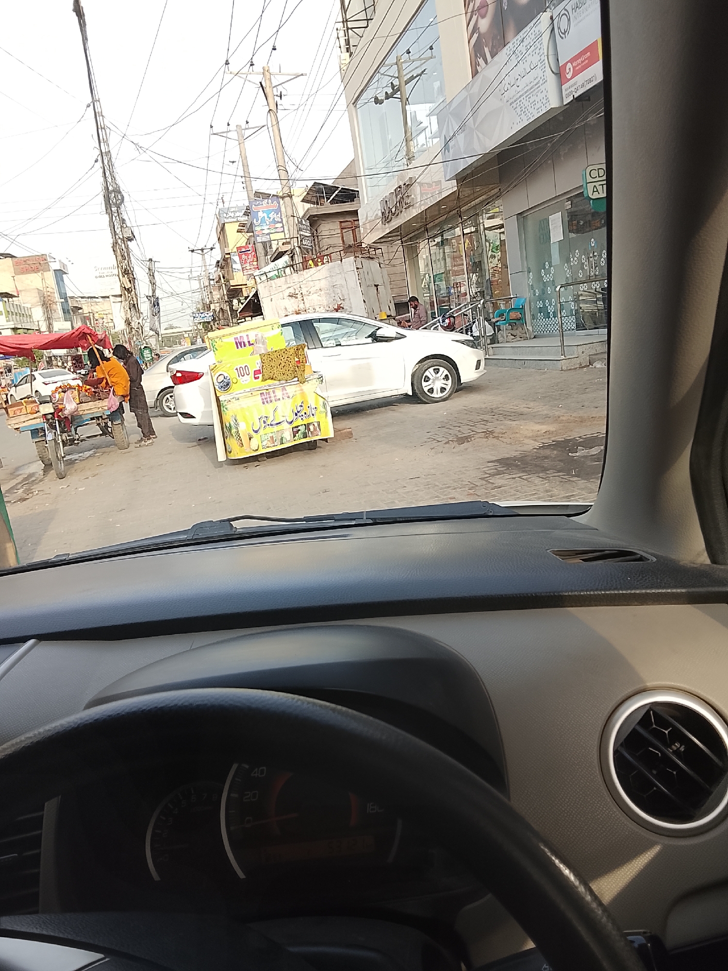 The image shows a view from inside a car, seemingly stationary, looking out onto a street scene in Gulgasht Colony, Pakistan. A white car is parked on the roadside. A fruit vendor's cart is positioned nearby, along with a juice vendor's stall displaying a sign in Urdu. Utility poles and power lines are visible overhead. A person wearing an orange shirt stands near the fruit vendor's cart. A multi-story building with glass windows and commercial signage can be seen in the background, including the name "Bank Alfalah Islami." The car's dashboard and steering wheel are prominently featured in the foreground.