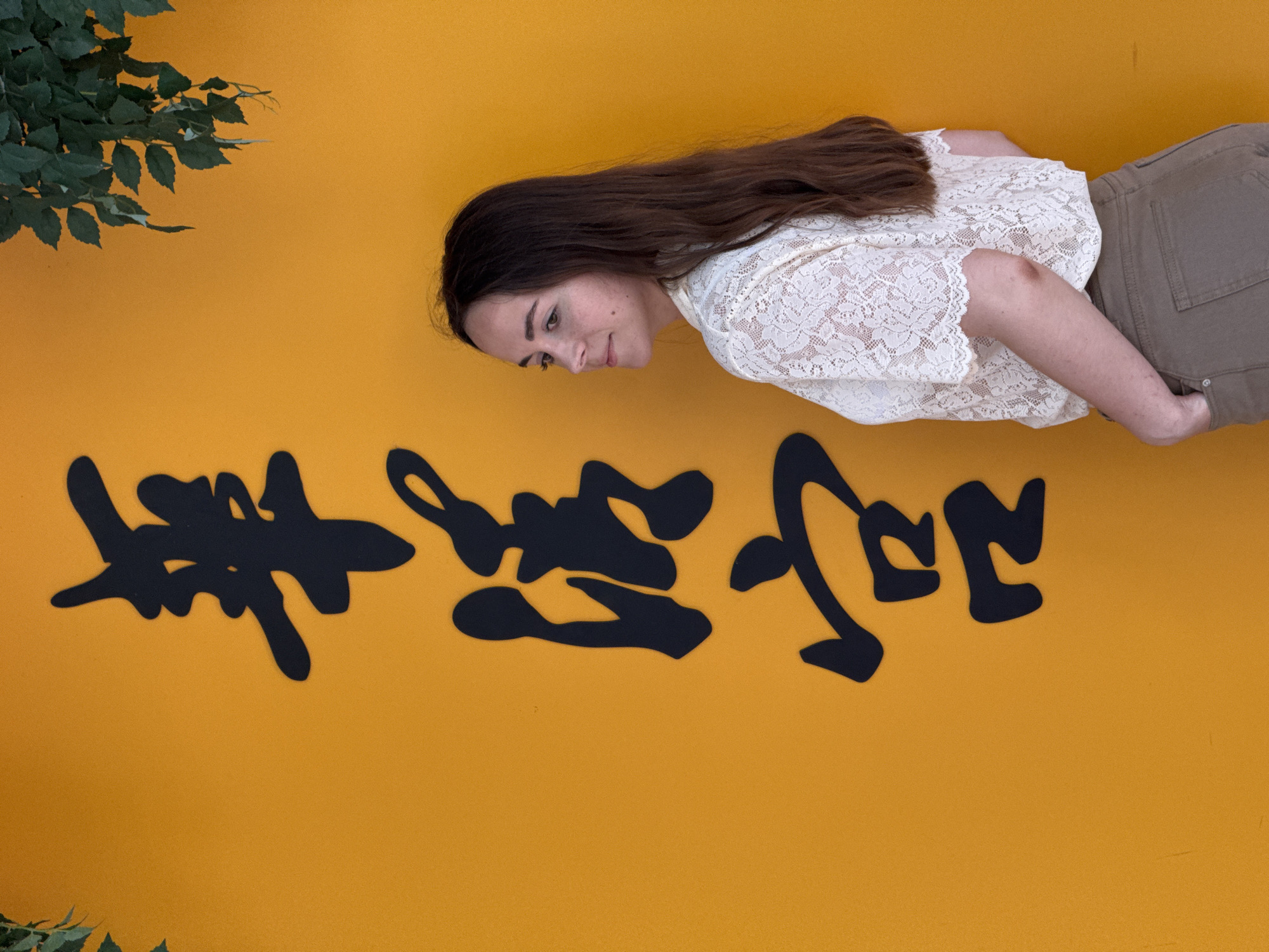 A young woman stands against a mustard yellow wall with large, black calligraphic characters running diagonally across it. The woman has long brown hair and is wearing a white lace top and olive-colored pants, with her right hand in her pocket. Green foliage is visible in the upper left corner of the frame. Based on the calligraphic characters and the location indicated as Lintong District, China, it's likely a decorative wall display.