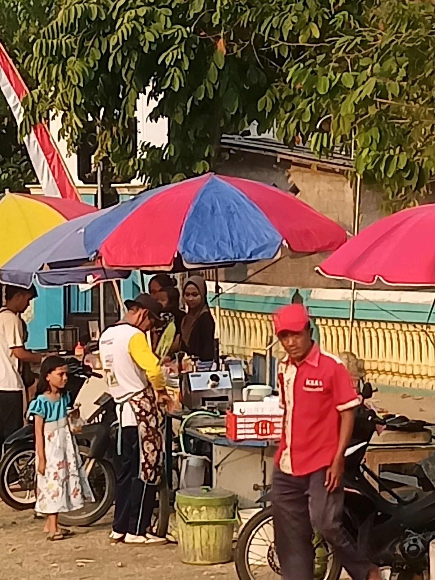 In Ketro, Indonesia, a street scene unfolds under the shade of colorful umbrellas. A man in a red shirt and cap, with "KLA" printed on his shirt, stands by a motorcycle, looking towards the viewer. Next to him, a vendor in a yellow and white shirt and a patterned apron prepares food at a cart. A young girl in a floral dress stands nearby, watching the vendor. Behind them, several people are gathered, including a woman in a headscarf and another woman in a black top. Red and blue umbrellas provide shade for a food stall. A motorcycle and a green trash bin are also visible in the scene, along with trees and buildings in the background. An Indonesian flag is partially visible in the upper left corner.