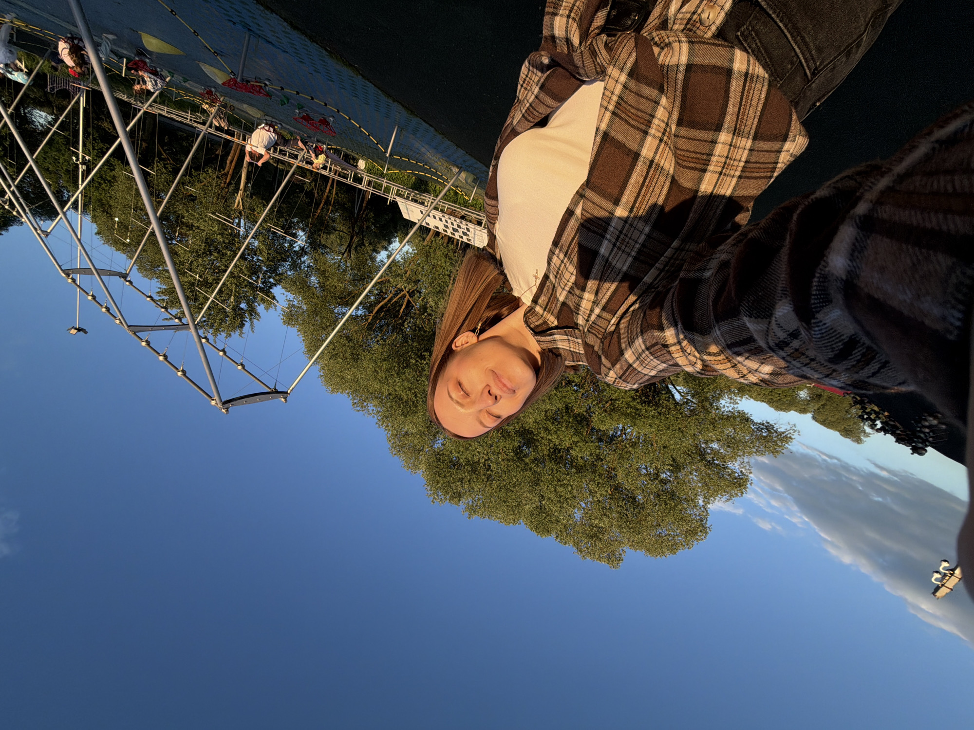 A woman is seen in the foreground, angled slightly downward in relation to the rest of the scene. She wears a white top under a brown and beige plaid flannel shirt. She has brown hair.
Behind her, the sky is visible. To the left, there is a tall metal structure reminiscent of an outdoor climbing frame. People can be seen on this climbing frame. Green trees fill the space around the climbing frame and beyond the woman. A section of what appears to be a blue surface, likely a playground or park floor, is visible above the climbing frame. The location is Sin'kovo, Russia.