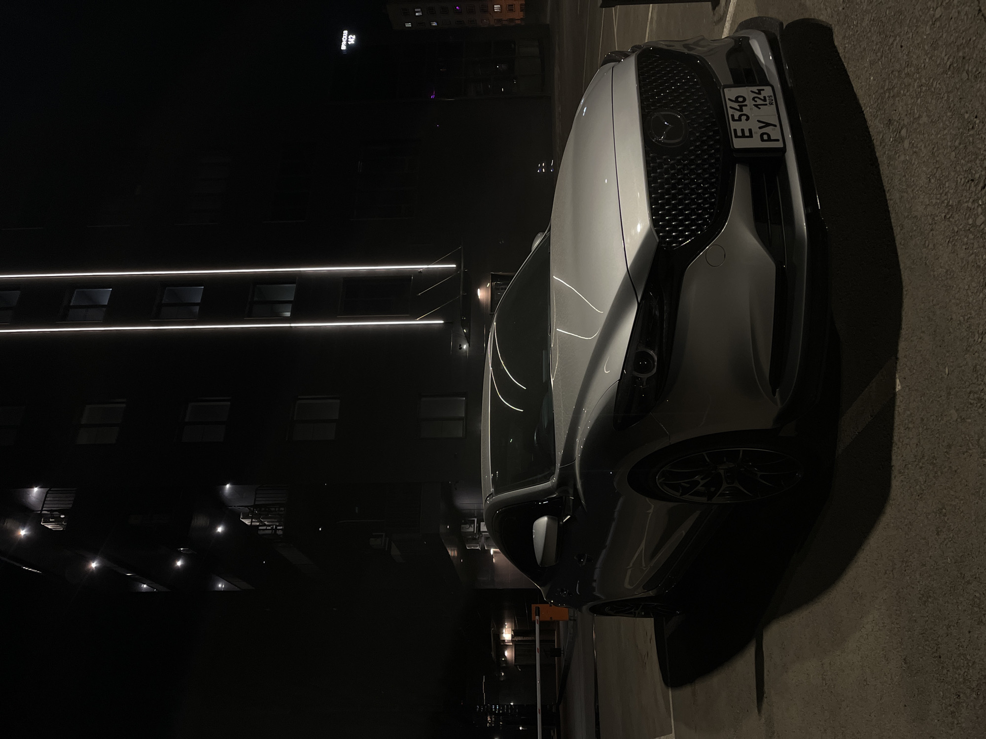 The image depicts a silver Mazda car parked on a dark paved surface, likely a parking lot or street, adjacent to a tall, dark building. The building features horizontal LED lighting that emphasizes its linear structure and windows arranged in a grid pattern. In the background, a taller building with lighted windows can be seen. The license plate on the car reads "E 546 PY 124 RUS", indicating it is registered in Russia. Based on the license plate information and the request, the location is Krasnoyarsk, Russia, though specific street address information is not provided in the image. No people are visible in the image.