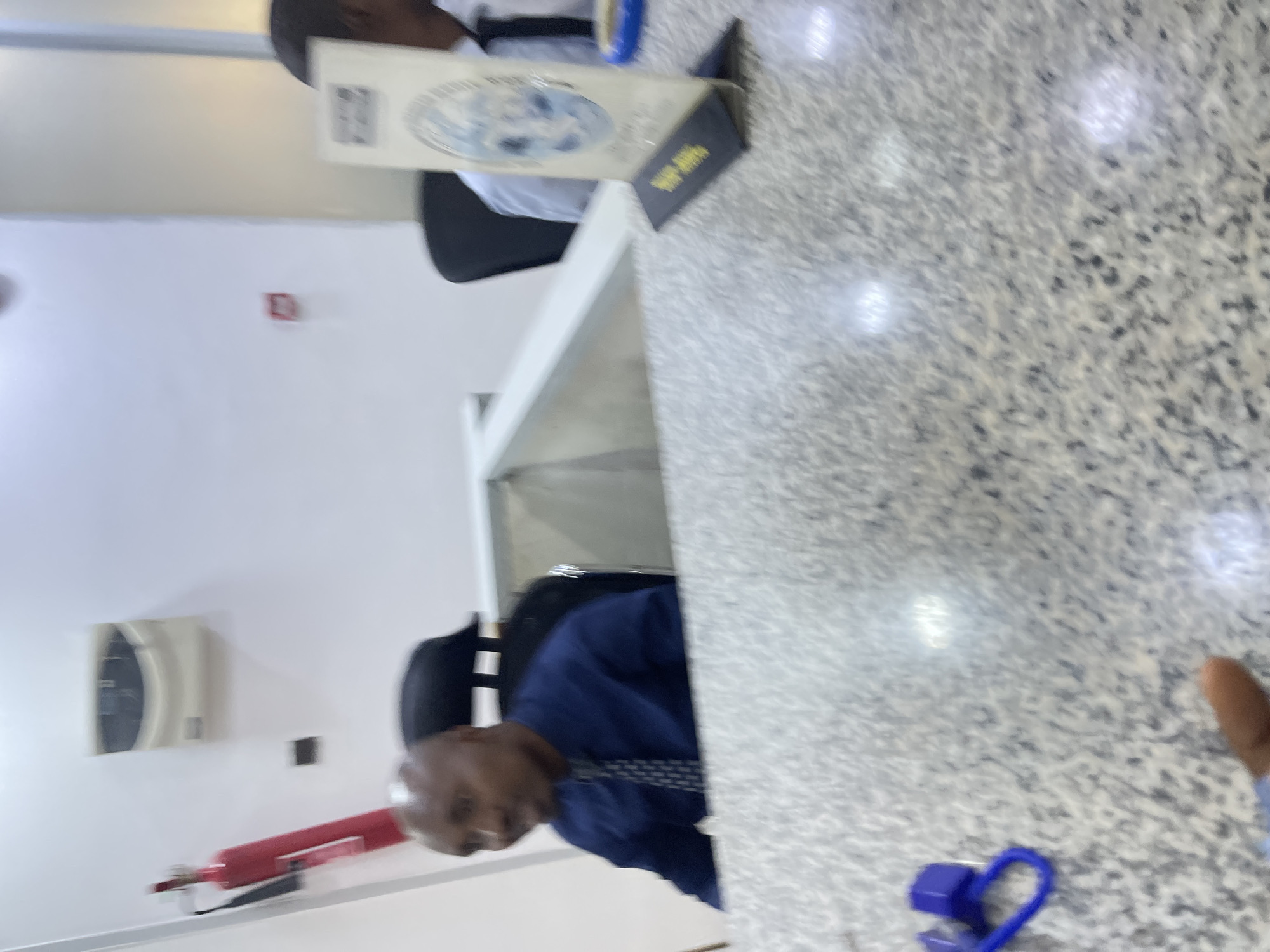 The image depicts an indoor scene, possibly a bank or a government office, in Jos, Nigeria. In the foreground, a speckled grey countertop is visible, with a blue cable and a purple object resting on it. In the mid-ground, a bald man wearing a blue collared shirt and a patterned tie is seated behind a partition, looking to his left. To his right, another person in a light blue shirt and a dark tie is partially visible. Behind the seated man, a black office chair is visible, and a red fire extinguisher is mounted on the wall to the left. A white unit is mounted on the wall above and behind the fire extinguisher. In the background, a sign with a blue and white design, possibly a service directory or informational display, is placed on the countertop. The environment appears to be a public service area, indicated by the arrangement of desks and the presence of official signage. The lighting suggests an indoor, possibly artificially lit, environment.