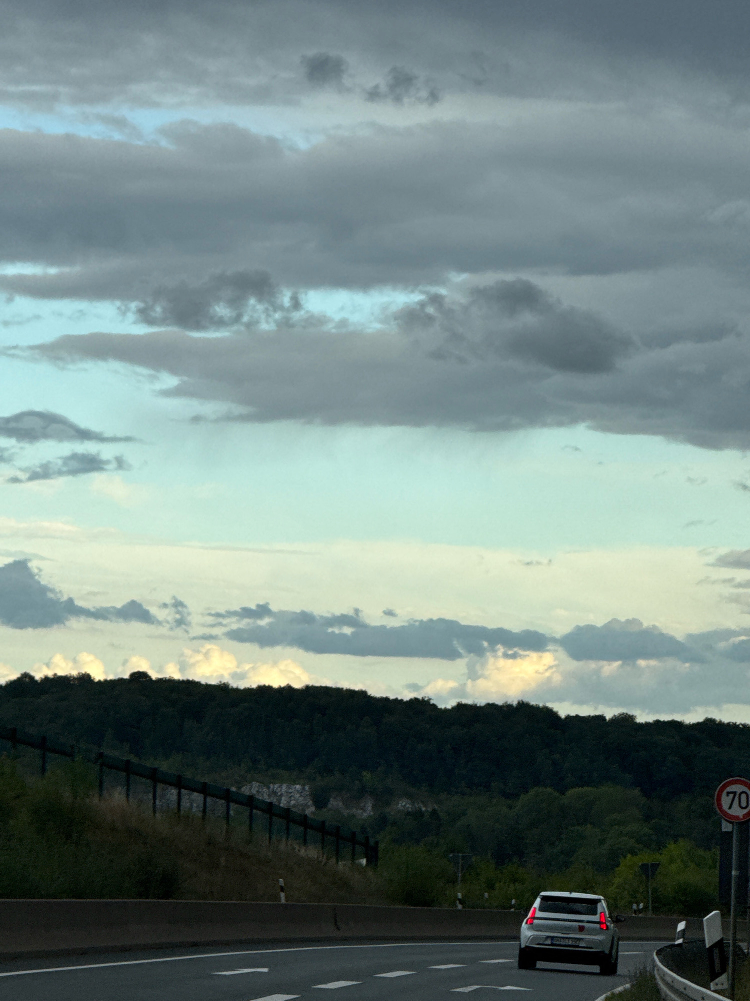 A white hatchback car drives on a highway in Bad Lauterberg im Harz, Germany, under a cloudy sky. The car, a white Fiat 500 with red taillights, is in the right lane of the highway. The highway is bordered by a concrete barrier on the left and a metal fence on the right, which separates the road from a wooded hillside. The hillside is covered in dense green trees, with a rocky outcropping visible in the middle. A speed limit sign indicating "70" is visible on the right side of the road. The sky is a mix of light blue and dark gray clouds, with patches of sunlight breaking through. The overall impression is a typical highway scene on an overcast day.