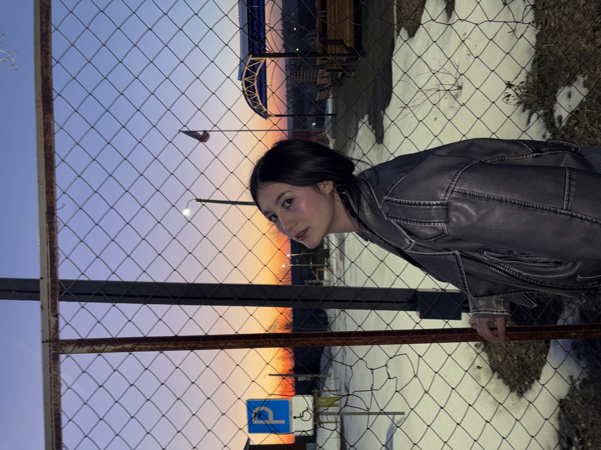 A woman in a gray leather jacket stands behind a chain-link fence. The fence partially obscures the view of a colorful sunset. Visible behind the fence are a lamp post, a small flag, what appears to be a structure with a curved roof, trees, snow patches, and a blue parking sign with a white wheelchair symbol. The location is Фоки, Russia.