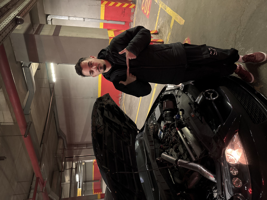 The image captures a young adult male standing next to a black Mazda vehicle with its hood open in an underground parking garage in Sofia, Bulgaria. The man, positioned to the right of the vehicle, possesses short dark hair, a trimmed mustache, and a goatee. He is dressed in a black puffer jacket over a dark hoodie, black cargo pants adorned with white graphics on the left leg, and reddish-brown sneakers with white soles. His hands are raised to chest level, with both index fingers pointed towards each other.
The black Mazda, likely a sedan, displays an illuminated driver-side headlight. The open hood reveals the engine compartment, showcasing an aftermarket red conical air filter and visible polished metallic intake piping among other engine components. The vehicle is parked on a concrete floor marked with yellow lines, indicating a designated parking space.
The environment consists of a concrete ceiling with exposed red fire suppression pipes and ventilation ducts. Fluorescent light fixtures are visible overhead. Concrete support pillars are present, some marked with yellow and red diagonal hazard stripes. A partially visible sign in the background, to the left, reads "ВХОД" (Entrance in Bulgarian), confirming the location as Sofia. A red symbol or text is partially visible on the floor to the right of the man.