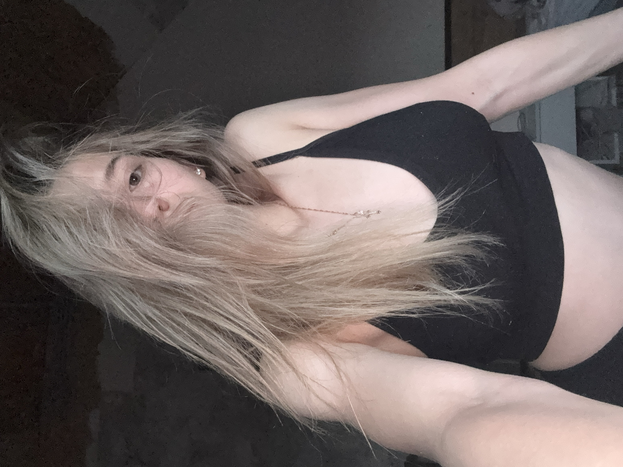 The media file features a close-up, upward-angled shot of a young individual, likely female, with long, light-colored (blonde or light brown) hair. The person is lying down, with their head slightly tilted and hair fanned out around their face and upper body. One eye is clearly visible, peeking through strands of hair, while the rest of the face is partially obscured. The visible eye appears to have a neutral or slightly contemplative expression.
The individual is wearing a simple black top, possibly a sports bra or crop top, which is visible across their chest and midriff. A delicate necklace with a small pendant hangs around their neck. One earlobe shows a small, silver-colored earring.
The background is uniformly dark and indistinct, suggesting an indoor setting with low or artificial lighting. There are no specific details in the background to indicate a precise location, other than it being an interior space. The overall impression is one of a relaxed, informal moment, possibly a self-portrait. No text is visible in the image. The image is provided in the context of Noviy, Russia.