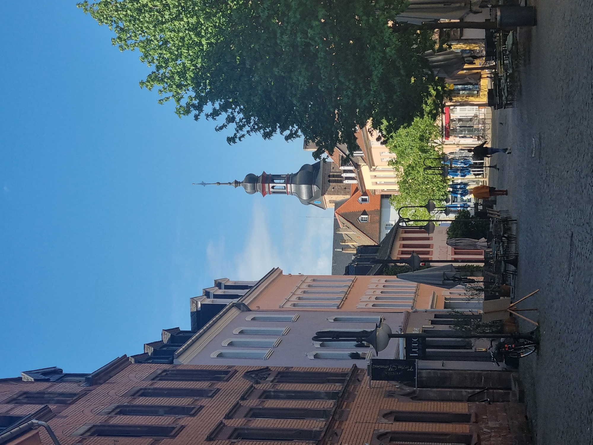 This image captures a vibrant urban scene on a clear, sunny day, likely a pedestrian street or town square in Saarbrücken, Germany. A brilliant blue sky, dotted with a few wispy clouds, dominates the upper portion of the frame, partially obscured by the lush, green canopy of a large tree on the left.
The left side of the scene is lined with multi-story buildings, showcasing varied architectural styles. The closest building features a reddish-brown brick facade with rectangular windows. Further down the street, buildings in shades of pale pink, light grey, and cream exhibit arched and narrow rectangular windows. Rising prominently above the rooftops in the background is a distinctive church spire, characterized by its tiered structure, black and red decorative elements, and a tall, slender pinnacle.
The right side of the image features a paved area, likely a pedestrian walkway or square, equipped with numerous outdoor cafe tables, chairs, and large sun umbrellas, most of which are closed. Streetlights, bicycle racks, and at least two bicycles are also visible. In the distance, two people are present: one person wearing a dark top and light pants walks away from the viewer, while another, clad in an orange or brown jacket, is also in the mid-ground.
The bright daylight indicates a clear day, possibly late morning or early afternoon, with shadows suggesting the sun is high in the sky. Visible text on black, pole-mounted signs includes "Trierer Straße" and "Restaurant," along with "Luuc" or "Lucc" on another sign. The atmosphere appears calm and peaceful, depicting an ordinary day in a European town.