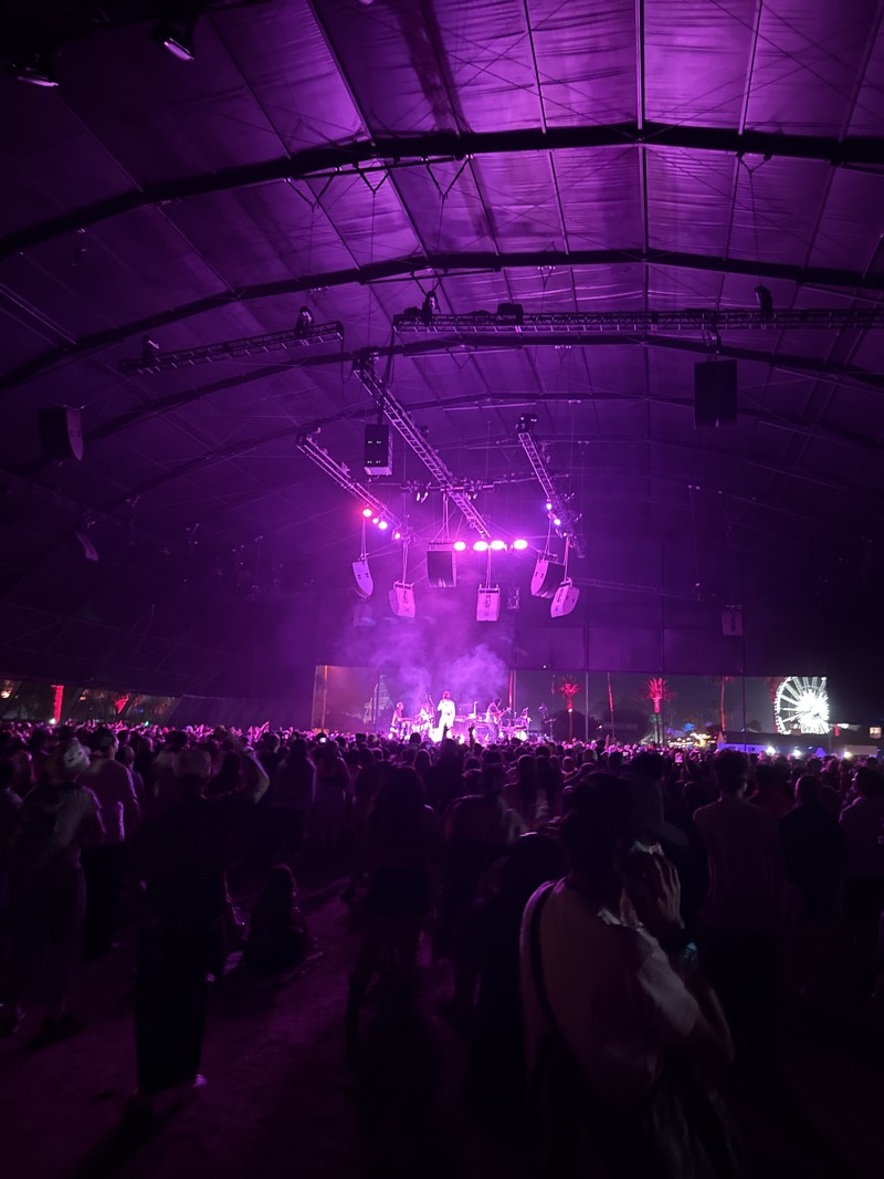 Captured indoors in Indio, United States, the image shows a music concert inside a large tent or dome-like structure, illuminated with purple lighting. The space is densely packed with people, facing the stage. On the stage, a band is performing, though their details are obscured by the distance and lighting. Stage lighting and sound equipment hang overhead. In the background, behind the stage, is a screen or backdrop, partially visible with palm tree imagery and a Ferris wheel. The roof of the tent is visible.
