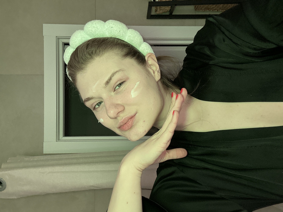 A woman with fair skin is shown in a selfie, wearing a green puffy headband and a black garment. Her hair is pulled back, and she has dabs of white cream on her forehead and cheek. Her fingernails are painted red. She poses with her hand near her face, which is tilted slightly. The background includes a white framed window, a pinkish-beige surface, and a black object partially visible in the upper right corner. The lighting appears soft and diffused.