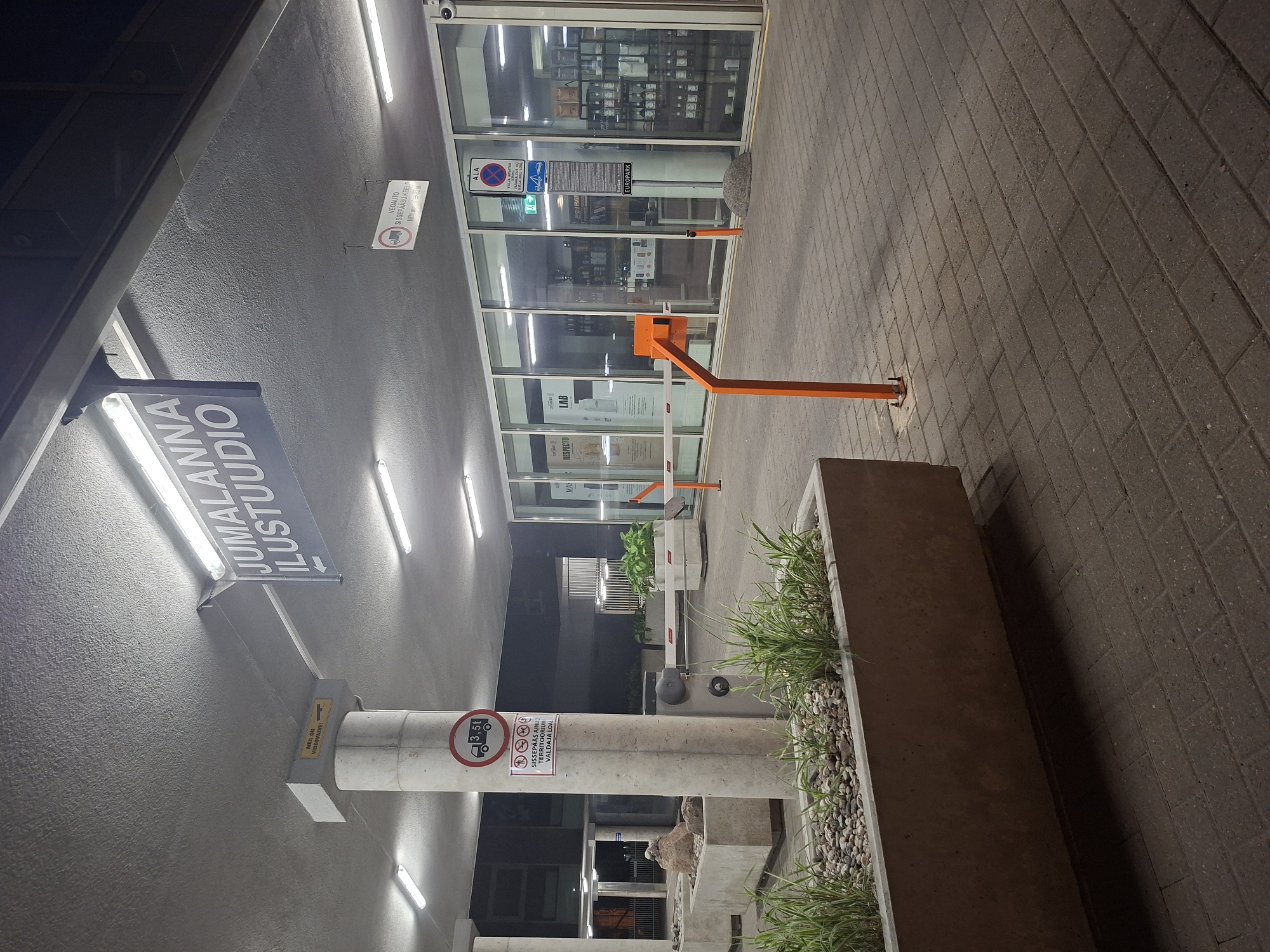 The image captures an exterior view of a building entrance at night, likely in Tallinn, Estonia. A prominent sign above the entrance reads "JUMALANNA ILUSTUUDIO" (Jumalanna Beauty Studio) with an arrow pointing left. To the left, a concrete pillar supports the overhang and displays several signs. One sign shows a truck icon with "3,5t" indicating a weight limit. Below this is a sign with multiple pictograms, including a truck, a car, and a person, with a red circle and slash through them, suggesting restrictions. Another small sign on the pillar reads "MEIL ON VIDEOVALVE!" (We have video surveillance!).
The entrance area features a paved walkway with planters containing grass and stones. A metal barrier, painted orange, is positioned to control vehicle access. The building facade consists of large glass windows, revealing the interior which appears to be a commercial establishment with shelves stocked with various products, possibly related to beauty or retail. A CCTV camera is visible mounted on the building's exterior near the entrance. The lighting suggests artificial illumination from the building's interior and overhead fixtures.