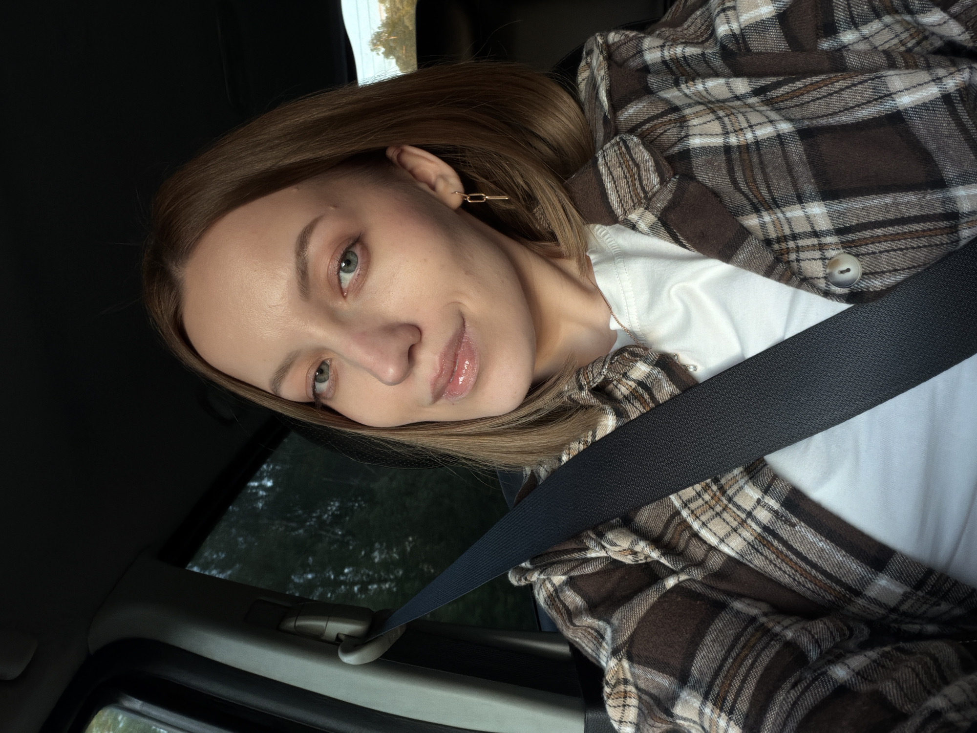 A woman is in a car, wearing a brown and white plaid shirt over a white t-shirt, and a seatbelt. She has light brown hair, fair skin, and green eyes. Her left ear has a gold chain-link earring. The background shows a window with greenery outside. There is no indication of the location being Rogachovo, Russia.