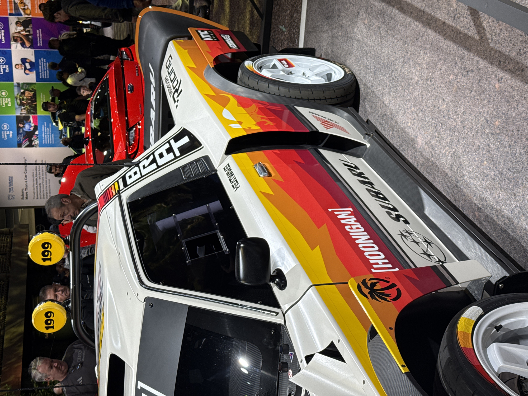 The image shows a modified white Subaru BRAT car on display, possibly at a car show or exhibition. The car features a custom orange and red paint job with "HOONIGAN" and "SUBARU" logos. It has white wheels, a roll cage with two yellow spotlights labeled "199," and is likely a rally or off-road-style build. People are standing around and observing the car. In the background, there's a red car and a promotional display with text and images, indicating the event may be related to the Subaru brand. The environment appears to be an indoor convention or exhibition center with carpeted floors. The location might be in New York City, United States of America, although this cannot be determined with certainty from the given image.