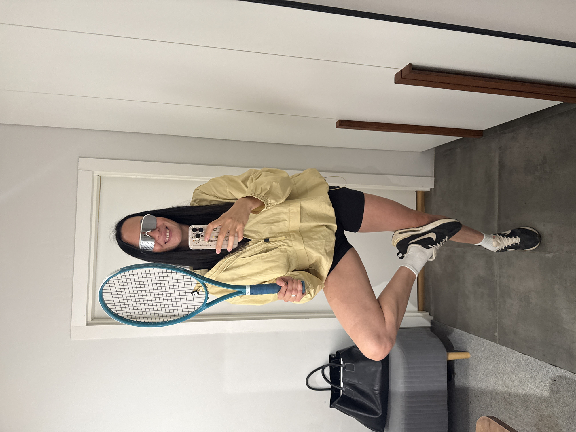A woman stands in a room, holding a tennis racket. She is wearing a light yellow jacket, black shorts, white socks, and black and white sneakers. She has on large, white-rimmed sunglasses and her hair is long and black. She is taking a selfie with her phone. In the background, there is a black handbag on a grey bench, a doorway, and a white closet. The flooring appears to be a mix of grey carpet and dark tile. The location is not specified as Chaikovsky, Russia.