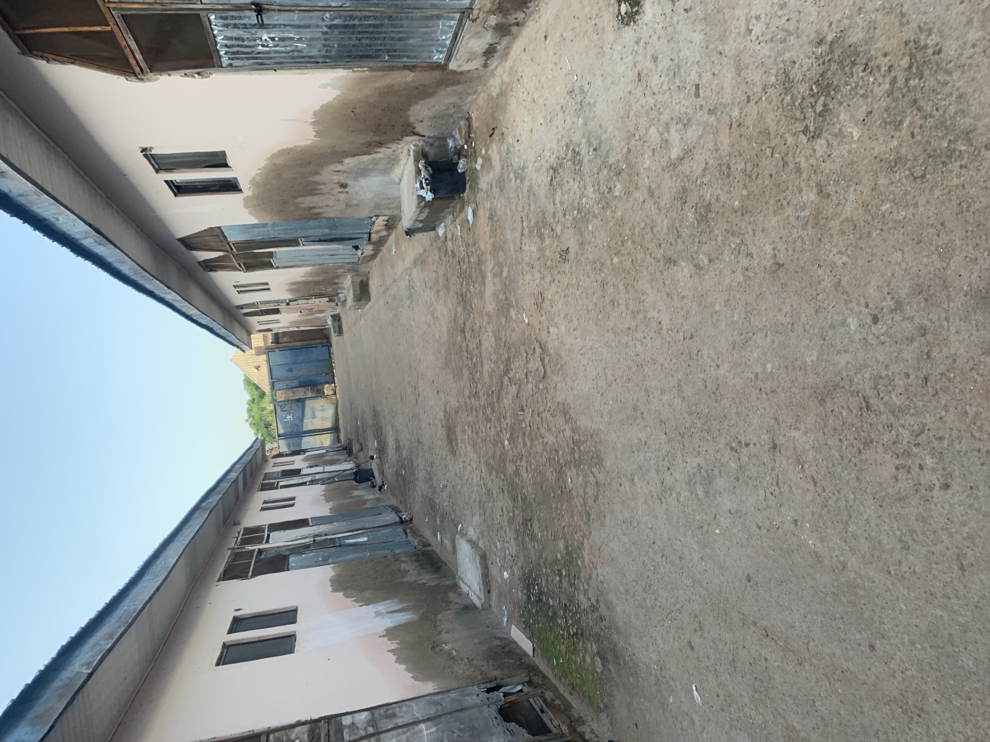 The image shows a view down a narrow alleyway between two rows of single-story buildings. The buildings appear to be made of concrete with metal roofs and corrugated metal doors and window coverings. The walls are painted a light pink and show signs of weathering and dirt. The ground is unpaved and appears to be a mixture of dirt and gravel, with scattered debris including plastic bags and white paper. There is a black trash bin overflowing with garbage on the right side of the alley. The sky is clear and blue. The scene suggests an urban or semi-urban environment, possibly residential or commercial in nature.