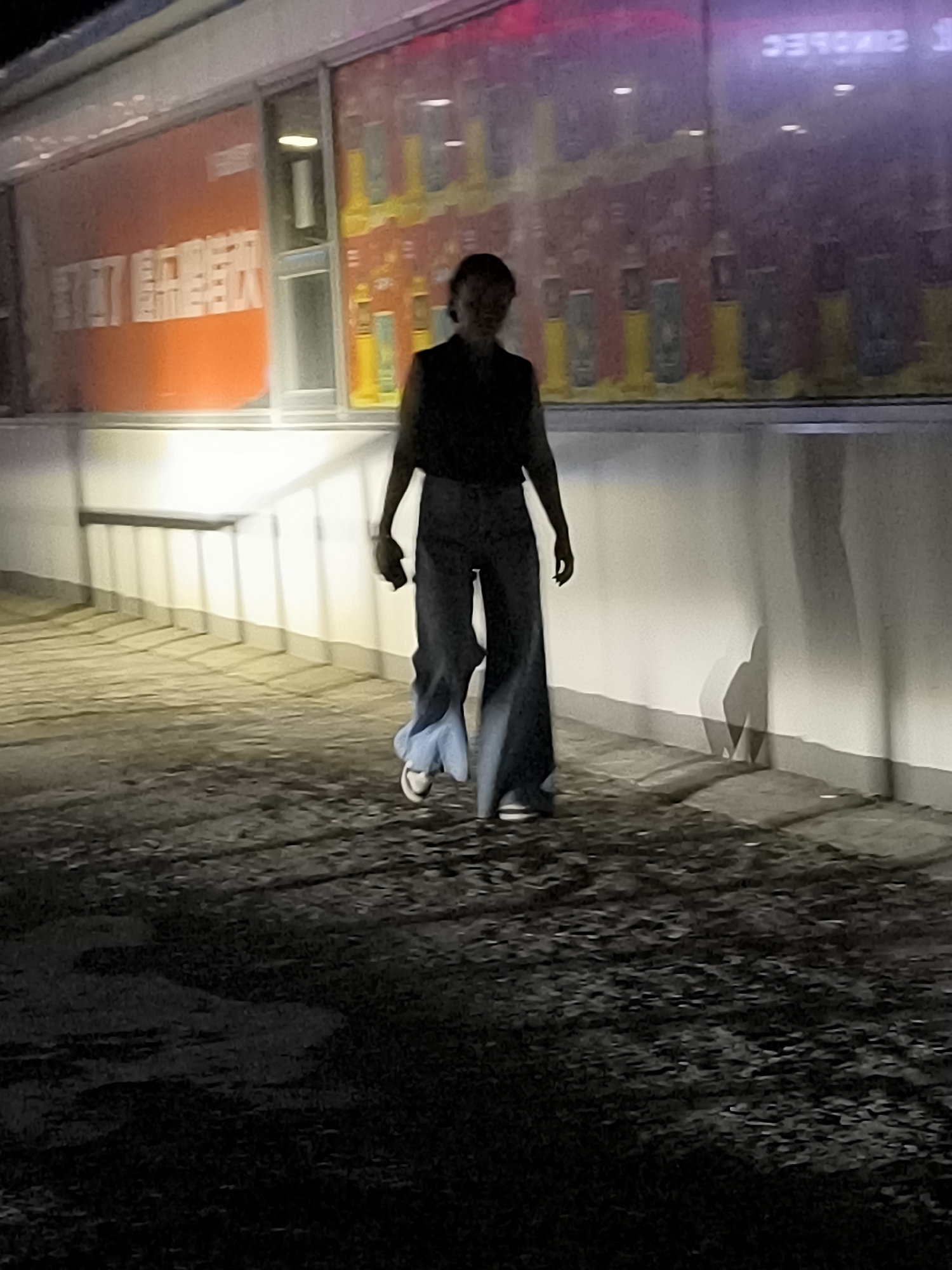A person walks down a paved pathway in front of a building. The building has a large window with an orange banner on the left side and a display of bottles on the right. The person is wearing a dark top and wide-leg jeans. The lighting suggests it is nighttime.