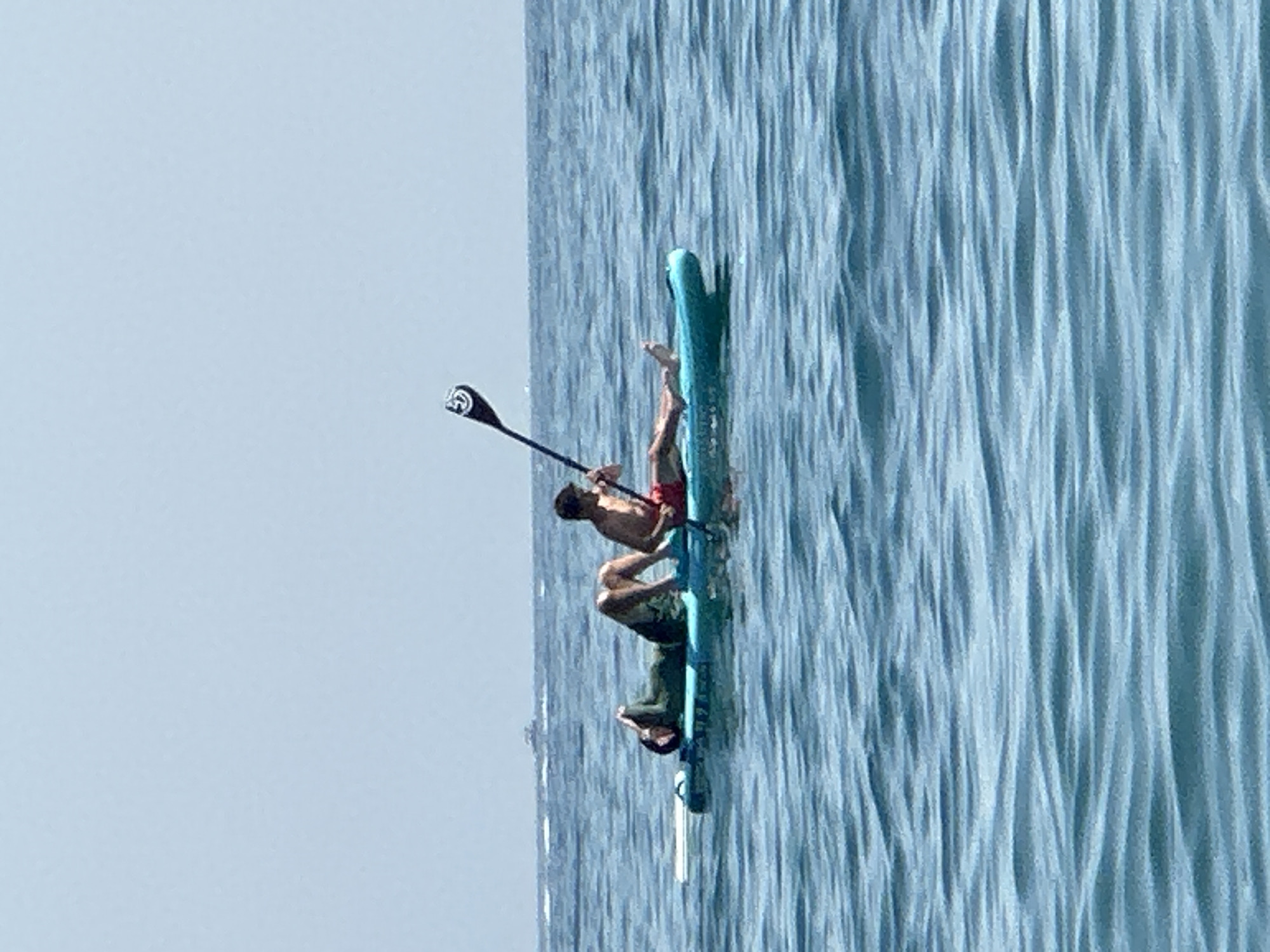 Two individuals are on a paddleboard in the calm, blue water of Boreti, Montenegro. One person, wearing red shorts, is sitting upright and holding a paddle, poised to move the board. The other individual, wearing a green long-sleeved shirt, is lying down on their back, resting. The paddleboard is turquoise and white. The background shows a clear, pale sky and the faint outline of a boat on the horizon.