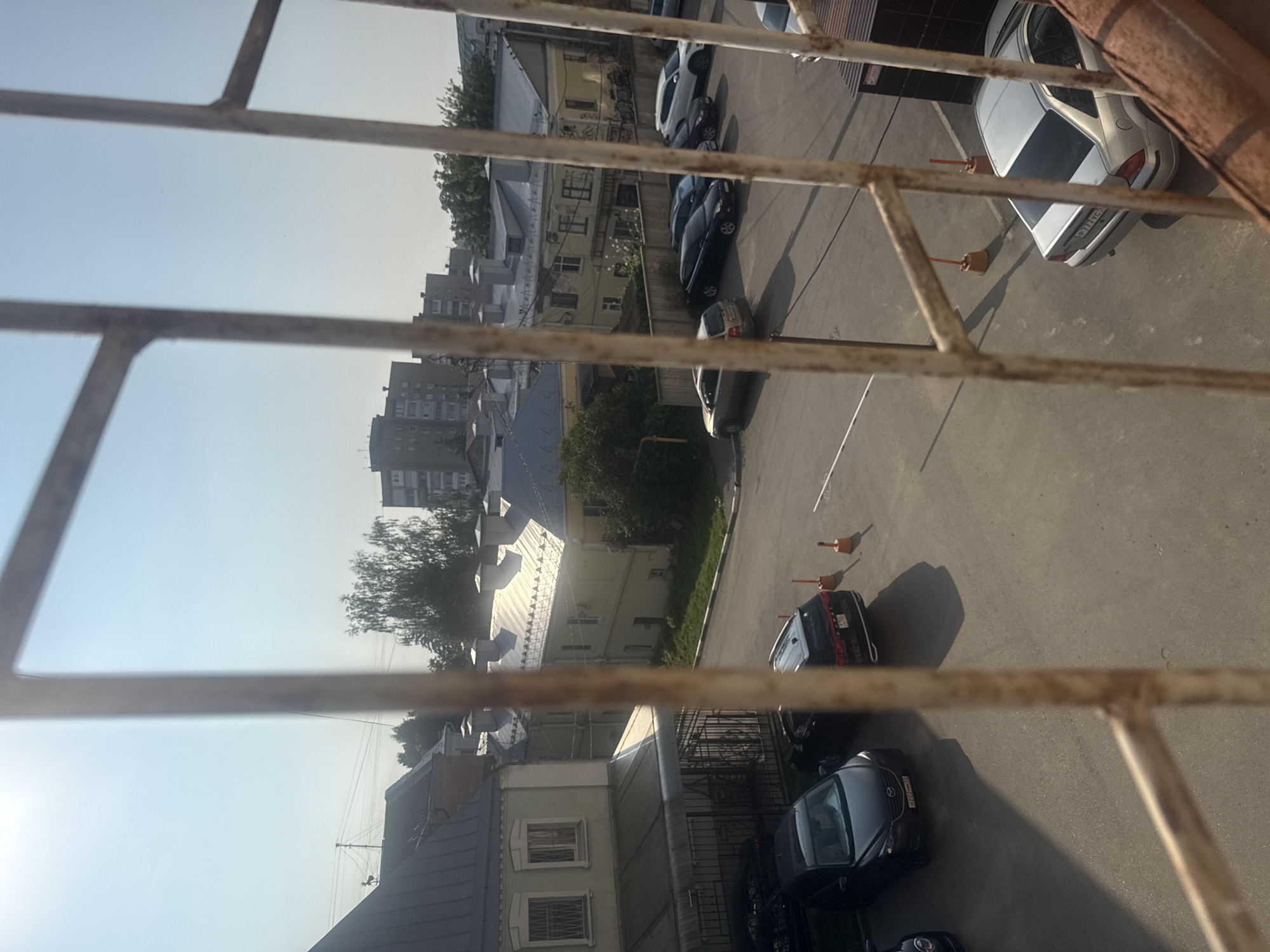The image depicts an outdoor parking lot in Oryol, Russia, viewed through a barred window. The foreground is dominated by the rusted metal bars of the window frame, which partially obscure the scene.
In the mid-ground, a paved parking lot is visible, with several cars parked. To the left, a dark-colored SUV is parked in front of a light-colored, multi-story building with a green facade and white trim. This building appears to have a covered walkway or entrance area. Further to the right, a black sedan is parked, and beyond it, a black SUV with its license plate visible. Two orange traffic cones are placed near the black SUV.
In the background, a series of residential buildings with light-colored facades and grey roofs are visible. Some of these buildings appear to be older, while one taller, modern apartment building stands further in the distance. Trees and overhead power lines are also present in the background. The sky is clear and blue, indicating a sunny day. The overall scene suggests a typical urban environment.