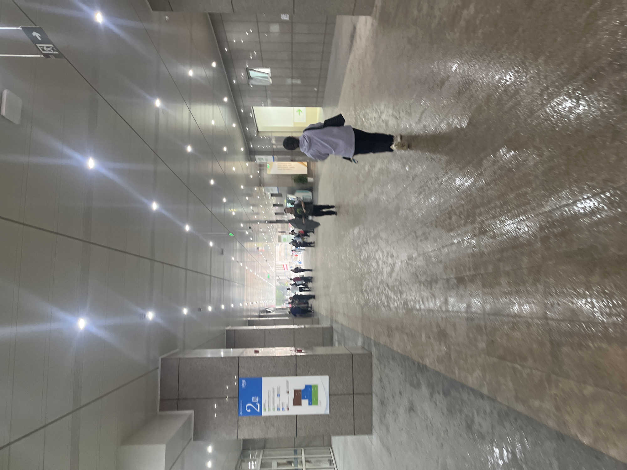The image depicts an indoor corridor or walkway with a polished, wet floor. Several individuals are visible, walking in various directions. On the left, a large pillar displays a blue and white sign with the number "2" and Chinese text. The ceiling is white, with numerous recessed lights. A sign with an arrow pointing to the right and Chinese characters is visible hanging from the ceiling on the right side of the image. The architecture suggests a modern public building, possibly an airport, train station, or convention center. The presence of water on the floor indicates it may have recently rained or been cleaned. The location in Yuzhong District, China, is a dense urban area known for its commercial and administrative functions.
