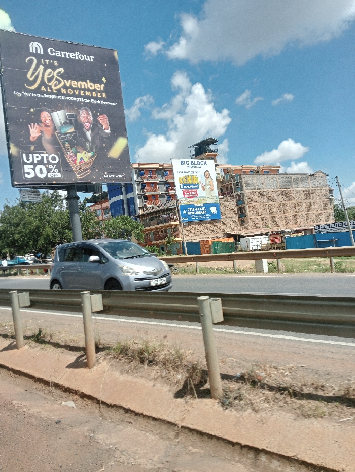 This daytime image captures a roadside scene, likely in Kenol, Kenya, under a bright blue sky with scattered white clouds. A silver hatchback car, possibly a Toyota Ractis or Verso S, is seen driving on a multi-lane road, partially obscured by metal guard rails that separate the roadway from a dirt and grass median.
Dominating the left side of the frame is a large Carrefour billboard, prominently advertising "IT'S Yesvember ALL NOVEMBER," encouraging viewers to "Say 'Yes' to the BIGGEST DISCOUNTS this Black November" with "UPTO 50% OFF" available from "NOV 1 - 30." The billboard features two excited individuals, a woman and a man, cheering alongside a shopping cart overflowing with various products, conveying a sense of enthusiasm for the promotions.
Further in the background, to the right, a smaller billboard advertises "BIG BLOCK PROPERTIES," showcasing "HOMES FOR SALE" including "RENDOR (MANG’U)" at "Ksh 950k." Contact numbers "0712 444111" and "0722 488490" are also visible. Behind this billboard, several multi-story buildings, some appearing to be under construction or renovation with exposed brickwork, line the horizon. To the far right, a small sign for "RAS AUTO GARA" (likely "GARAGE") can be faintly seen. The visible local advertisements and phone numbers align with the provided location context of Kenol, Kenya.