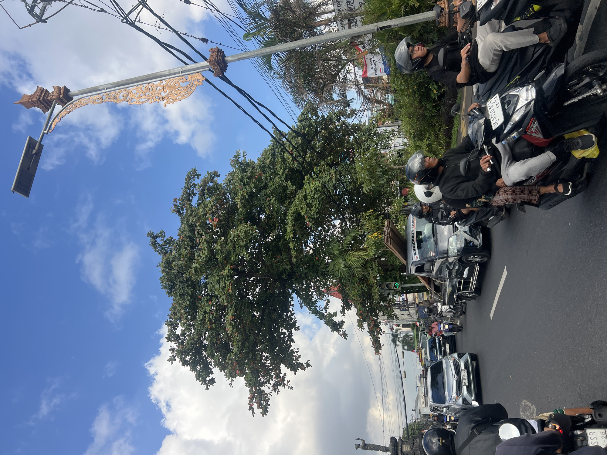 A medium shot captures a traffic scene in Denpasar, Indonesia, under a bright, partly cloudy blue sky. A large, leafy tree dominates the left side of the frame, its branches extending over a busy street filled with vehicles. On the road, several motorcycles navigate through traffic, with riders wearing helmets. Some motorcycles carry multiple passengers. A silver car with a license plate visible is also present, along with a black pickup truck carrying lumber. A decorative lamppost, characteristic of Indonesian street decor, stands on the right side of the road, adorned with ornate carvings. Electrical wires crisscross above the street. In the background, buildings and lush green vegetation are visible, suggesting a tropical urban environment.