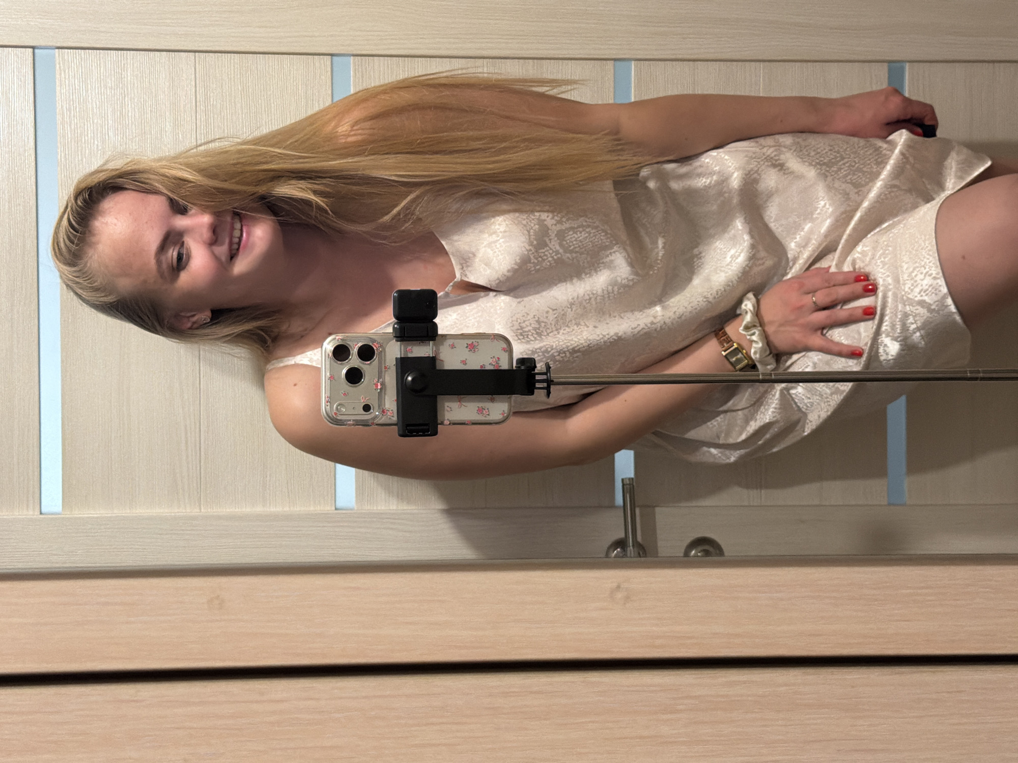 A woman with long blonde hair is smiling while taking a selfie using a phone attached to a selfie stick in front of a mirror. She is wearing a light-colored, patterned satin nightgown. She has a watch and a scrunchie on one wrist and red nail polish. The reflection shows a light-colored paneled door behind her. The location cannot be determined from the given image.